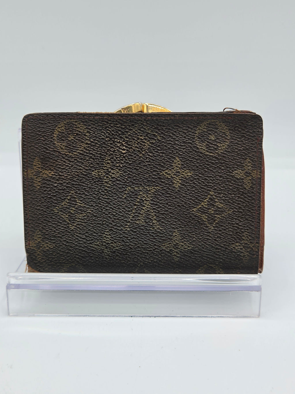 A Louis Vuitton monogram canvas wallet with a gold-tone clasp, shown standing upright next to a clear acrylic block against a plain white background.