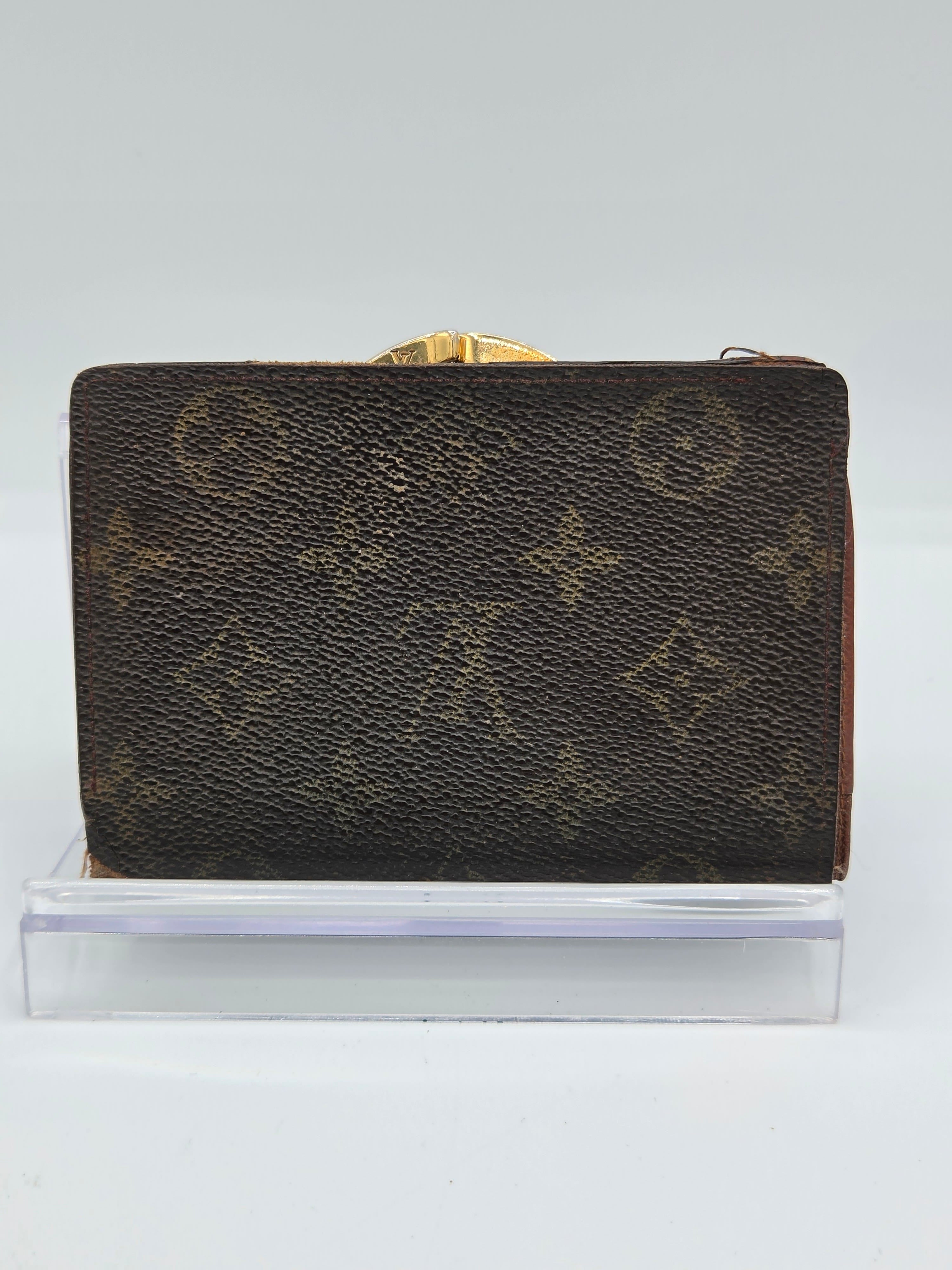 A Louis Vuitton monogram canvas wallet with a gold-tone clasp, shown standing upright next to a clear acrylic block against a plain white background.