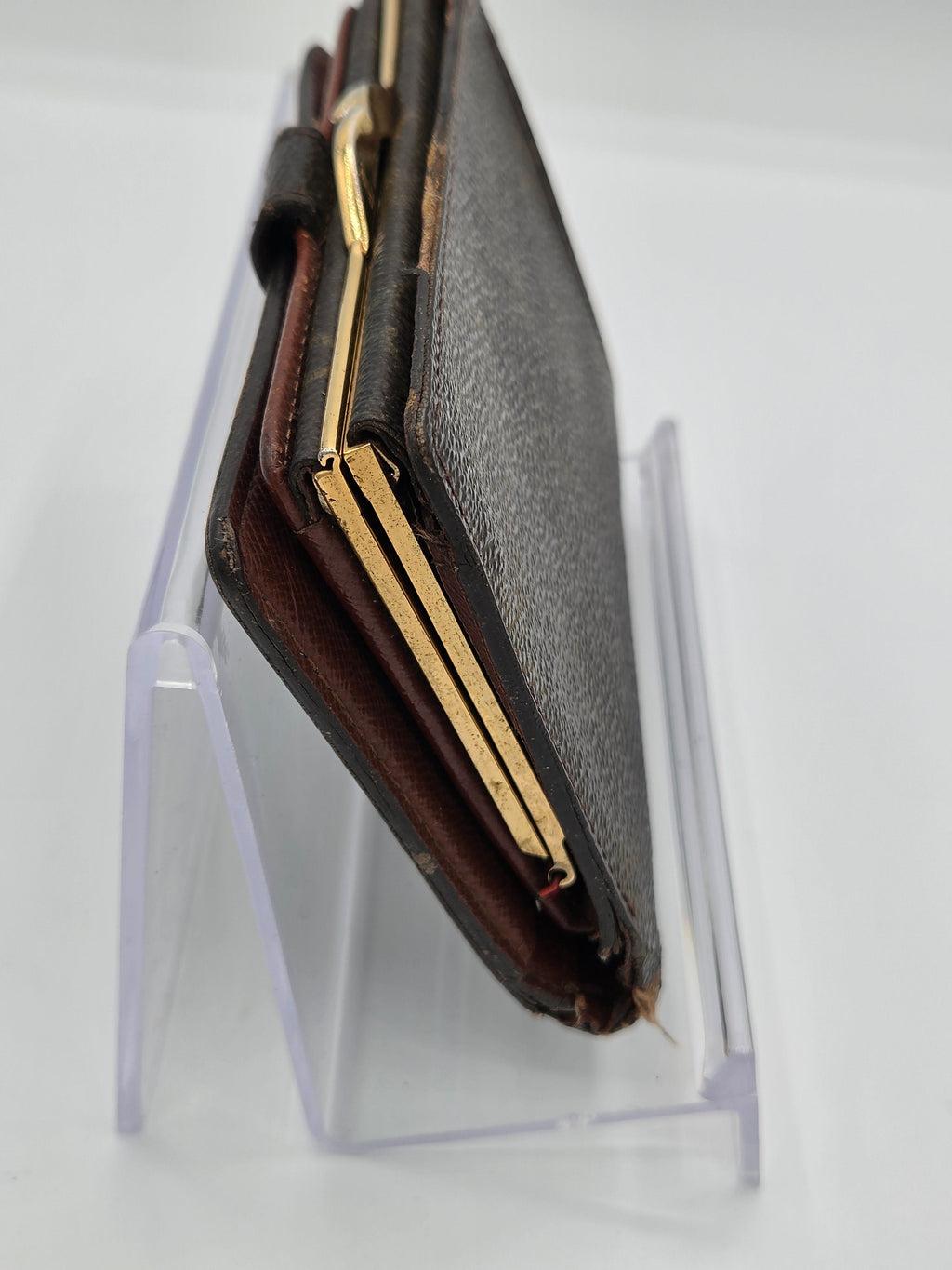 A dark brown textured leather wallet with gold-toned metal edges, resting on a clear acrylic display stand against a white background.