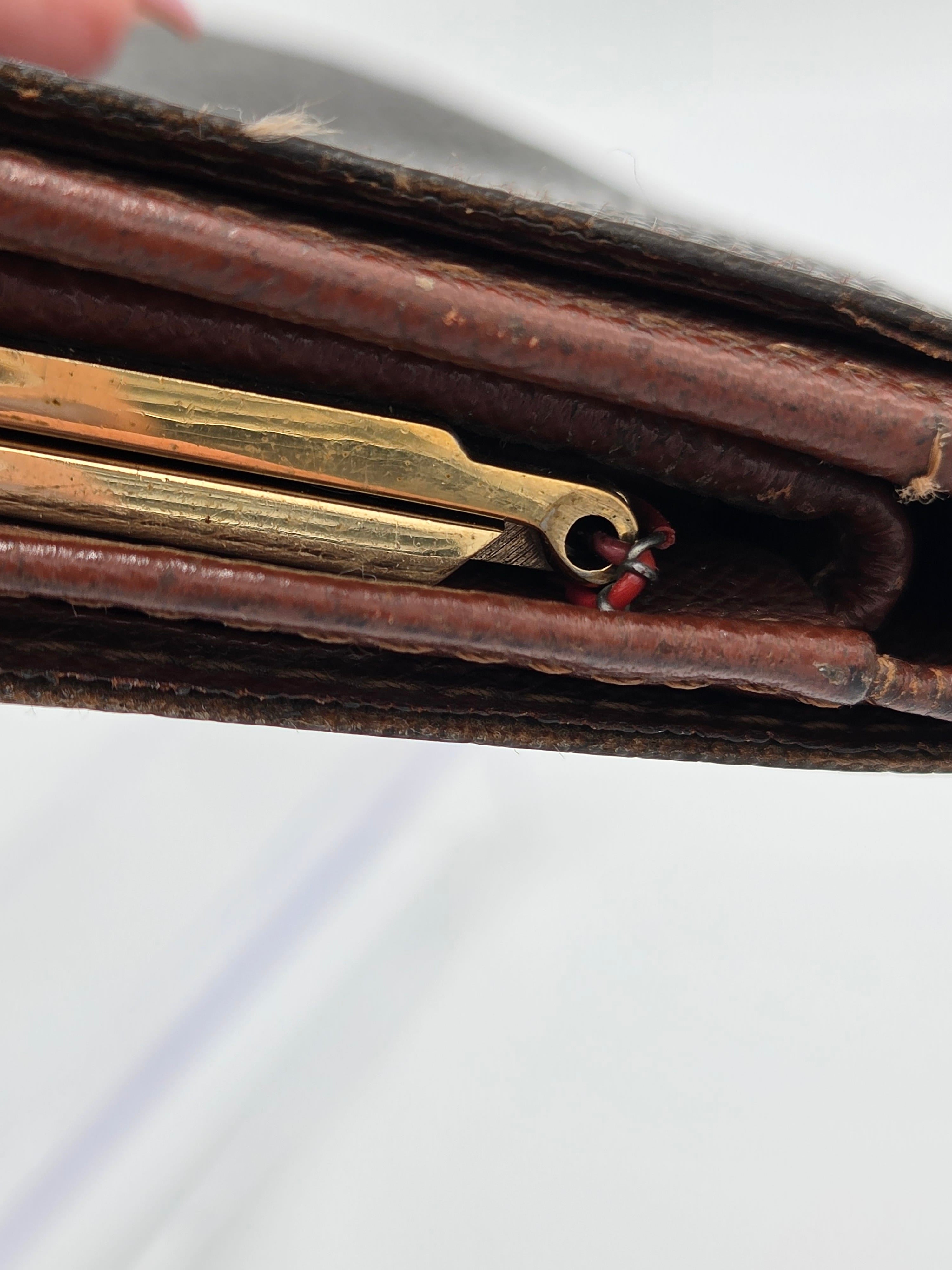 Close-up of a vintage brown leather case or wallet showing a brass-colored metal tool or charm hanging from the zipper area, with a white background.