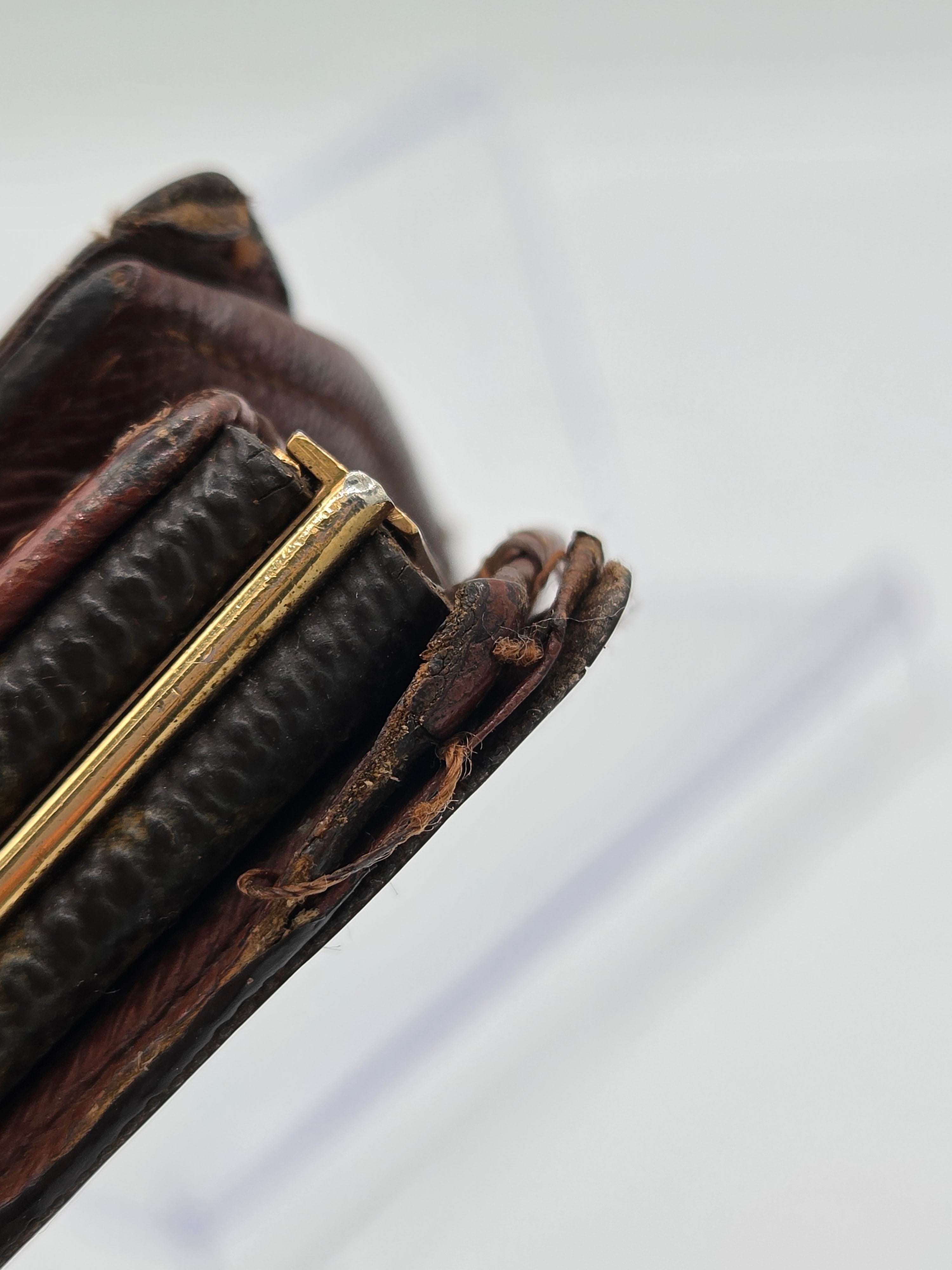 A close-up of a vintage dark brown leather wallet or clutch with a textured black leather band and a gold-tone metal frame, set against a soft, out-of-focus light gray background.