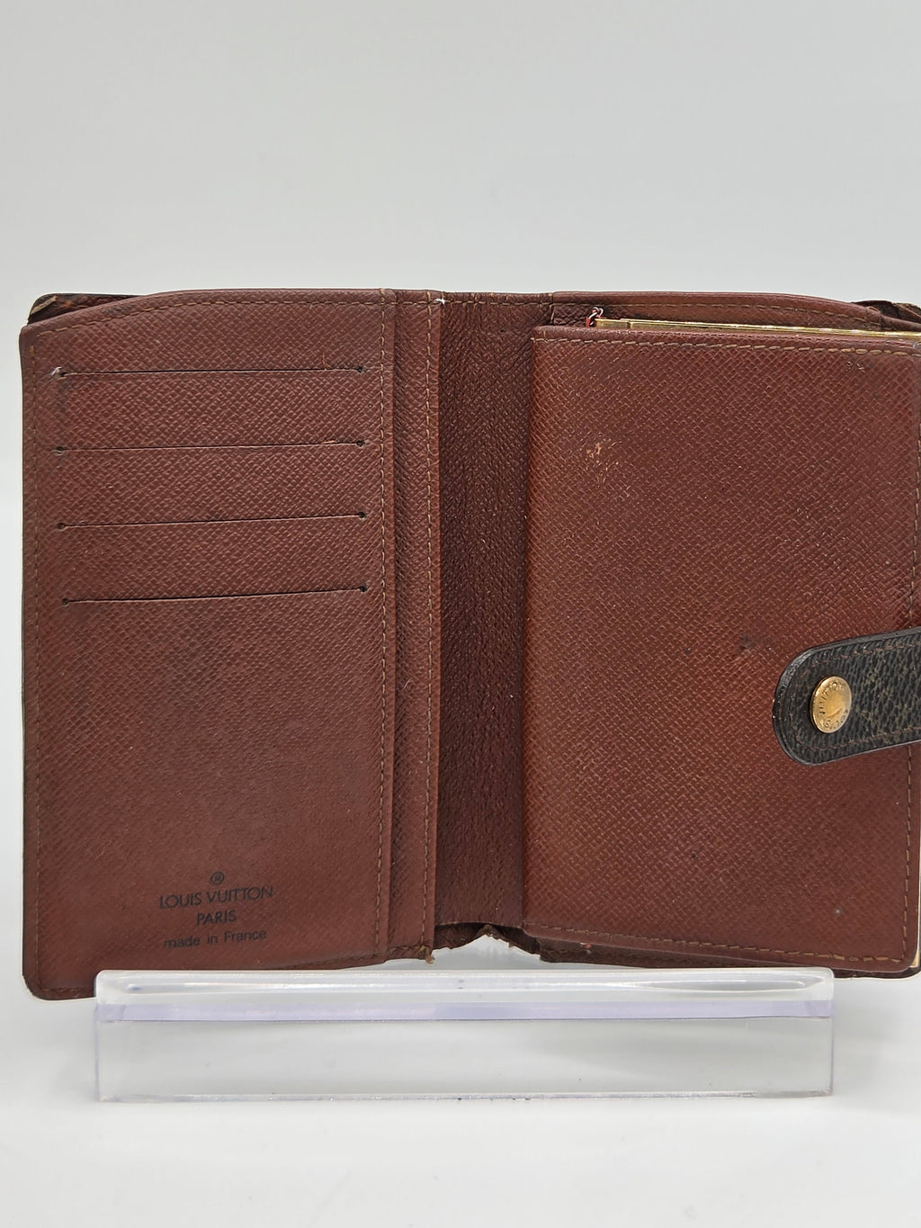 A brown textured leather wallet shown open, revealing multiple card slots and a dark brown leather tab with a gold-tone snap button. A clear plastic sleeve is positioned to the right of the wallet against a plain white background.