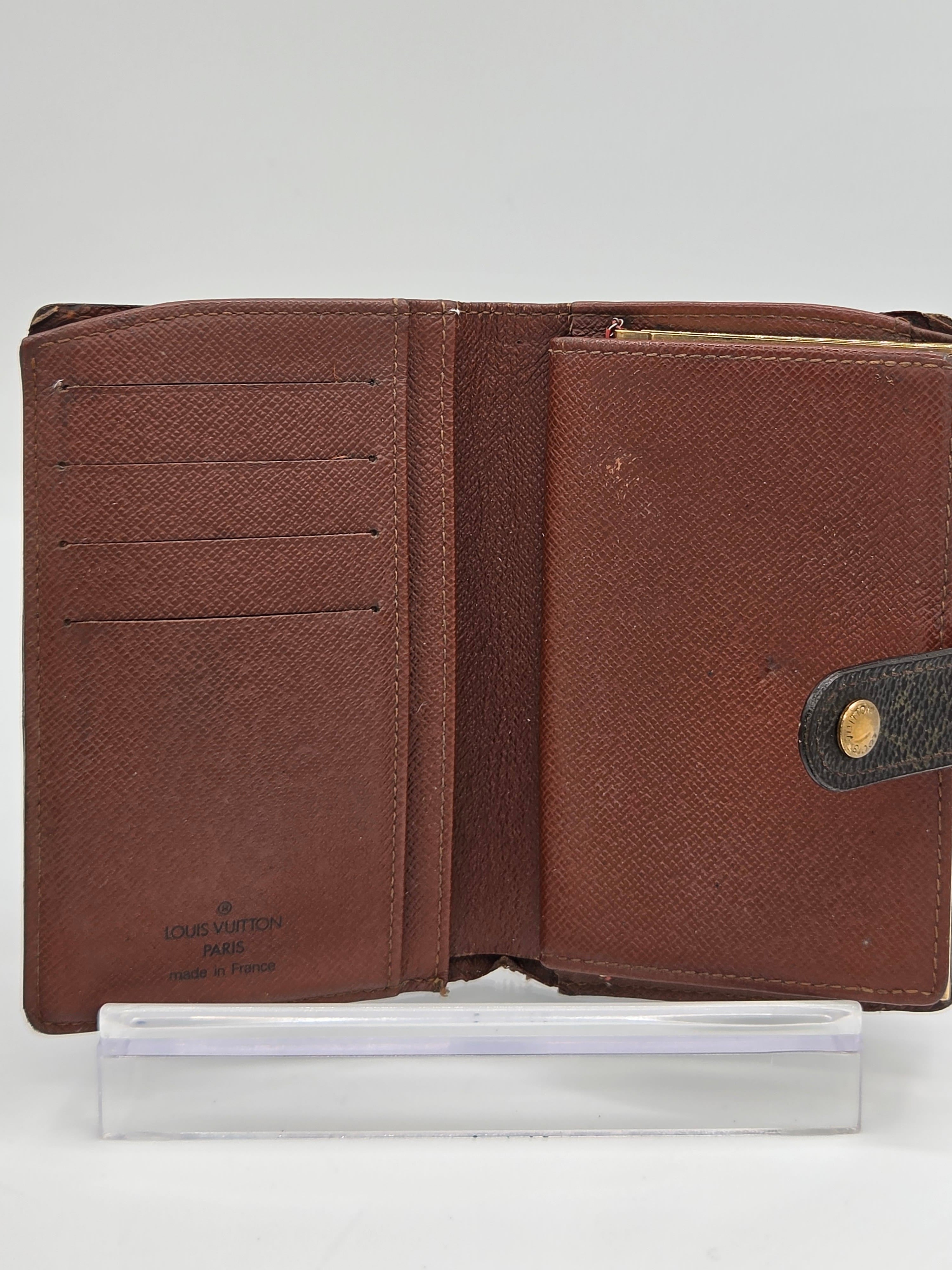 A brown textured leather wallet shown open, revealing multiple card slots and a dark brown leather tab with a gold-tone snap button. A clear plastic sleeve is positioned to the right of the wallet against a plain white background.