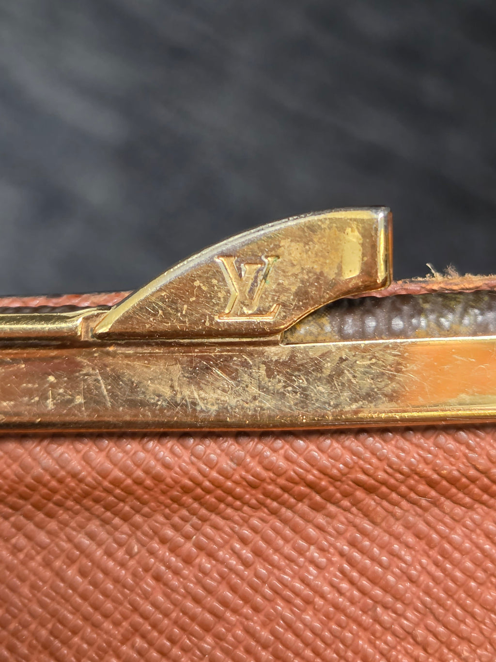 Close-up of a Louis Vuitton monogram zipper pull in gold-tone metal, showing the LV logo, next to a textured brown leather or coated canvas material.