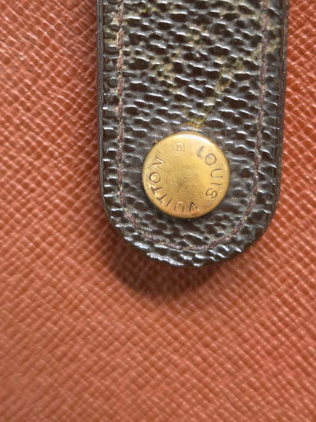 Close-up of a Louis Vuitton textured leather strap with a round gold-tone metal stud engraved with the brand name, set against a brown textured background.