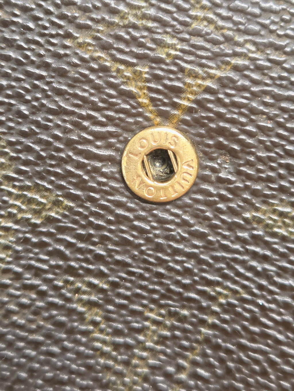 Close-up of a Louis Vuitton monogram canvas surface with a gold-tone metal snap button showing the brand name.