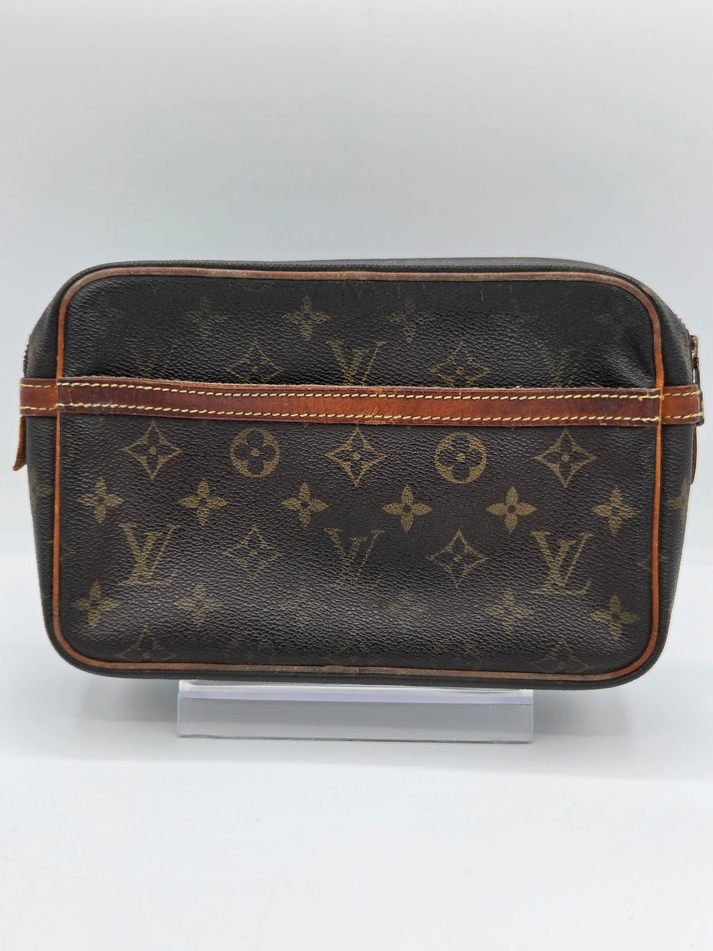 A Louis Vuitton monogram canvas toiletry bag in dark brown with tan leather trim, displayed on a clear stand against a light gray background.