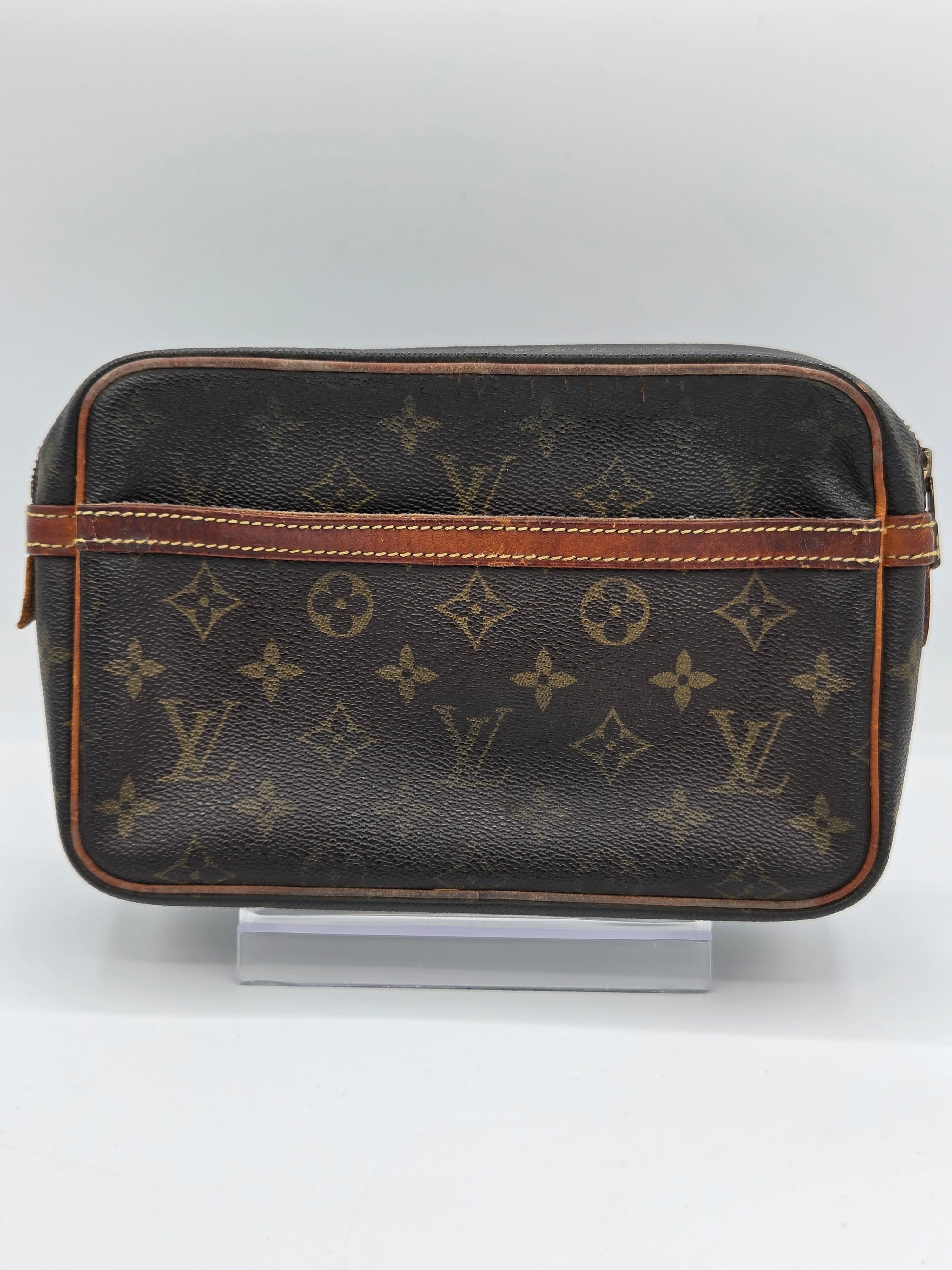 A Louis Vuitton monogram canvas toiletry bag in dark brown with tan leather trim, displayed on a clear stand against a light gray background.