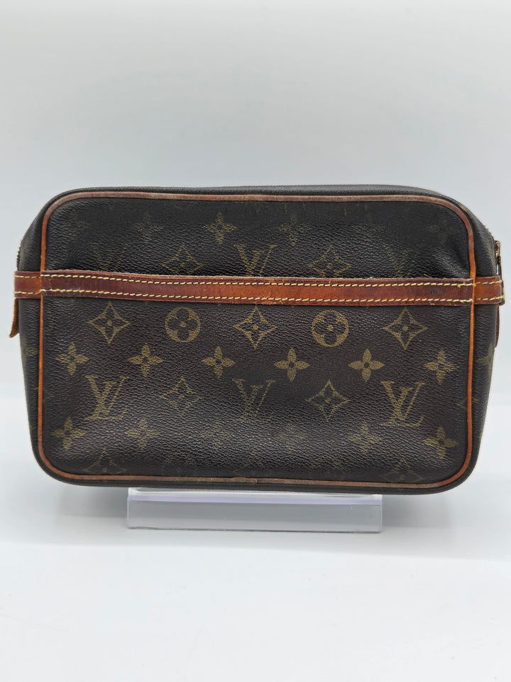 A Louis Vuitton monogram canvas toiletry bag in dark brown with tan leather trim, displayed on a clear stand against a light gray background.