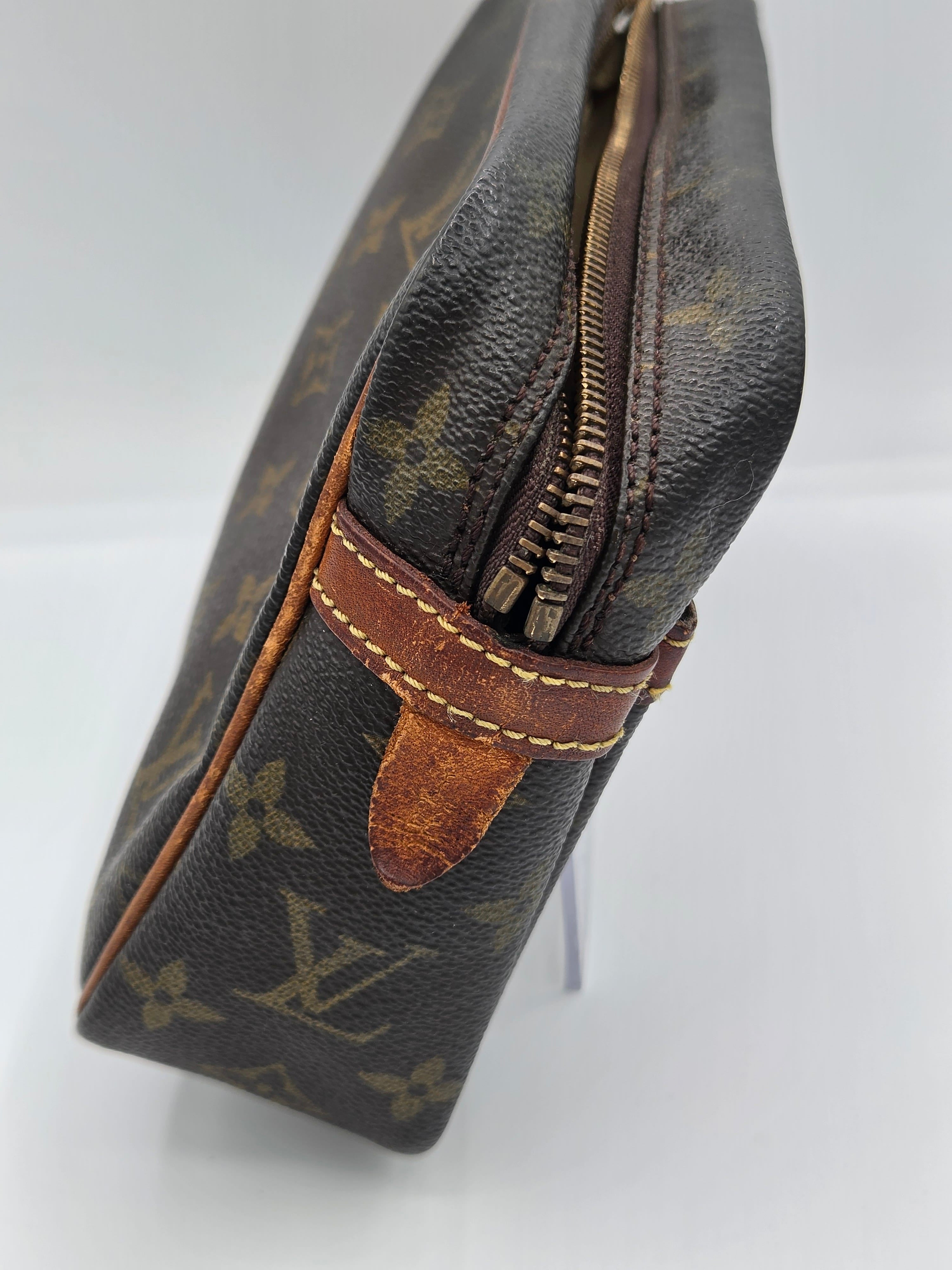 A close-up of a Louis Vuitton monogram canvas pouch or small bag, showing the dark brown coated canvas with tan monogram motifs, a brown leather zipper pull tab, and a metal zipper along the top edge.