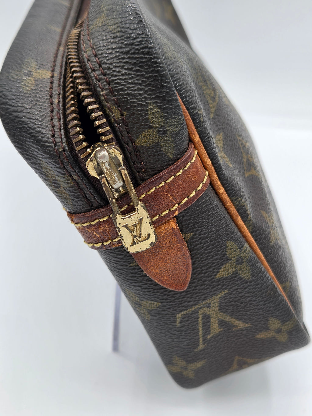 Close-up of a Louis Vuitton monogram canvas pouch or small bag, showing the dark brown coated canvas with tan monogram motifs, a brown leather trim, and a gold-tone zipper pull with an LV logo.