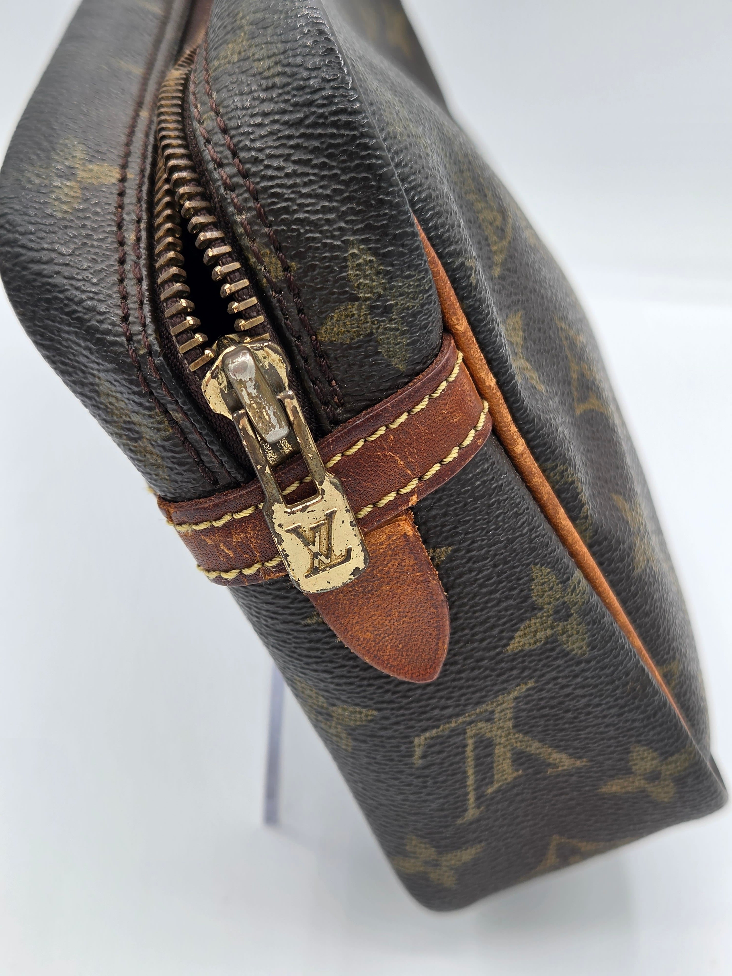 Close-up of a Louis Vuitton monogram canvas pouch or small bag, showing the dark brown coated canvas with tan monogram motifs, a brown leather trim, and a gold-tone zipper pull with an LV logo.