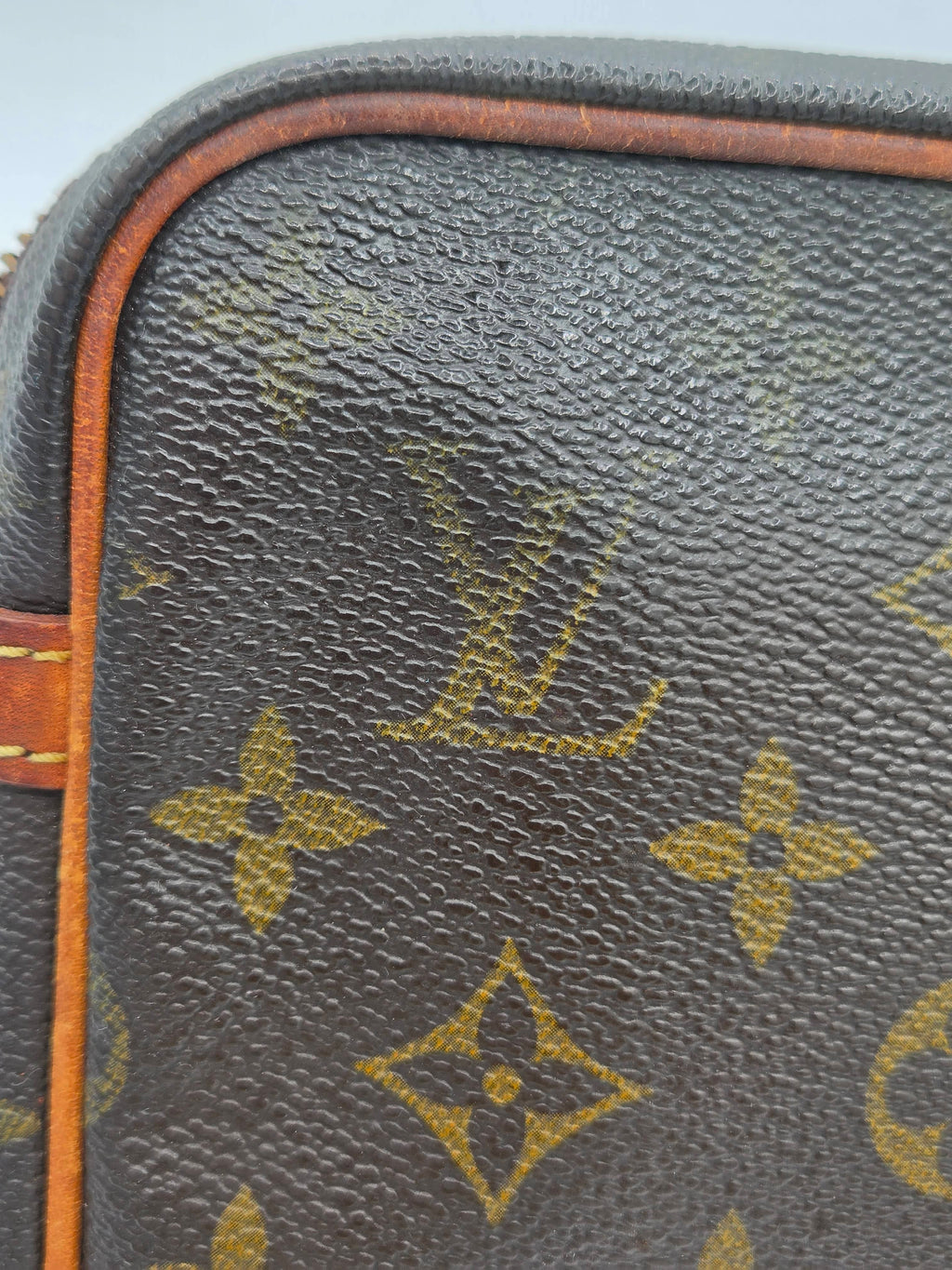 Close-up of a Louis Vuitton monogram canvas bag corner, showing the dark brown coated canvas with the classic tan/gold LV monogram pattern and a strip of orange-brown leather trim.
