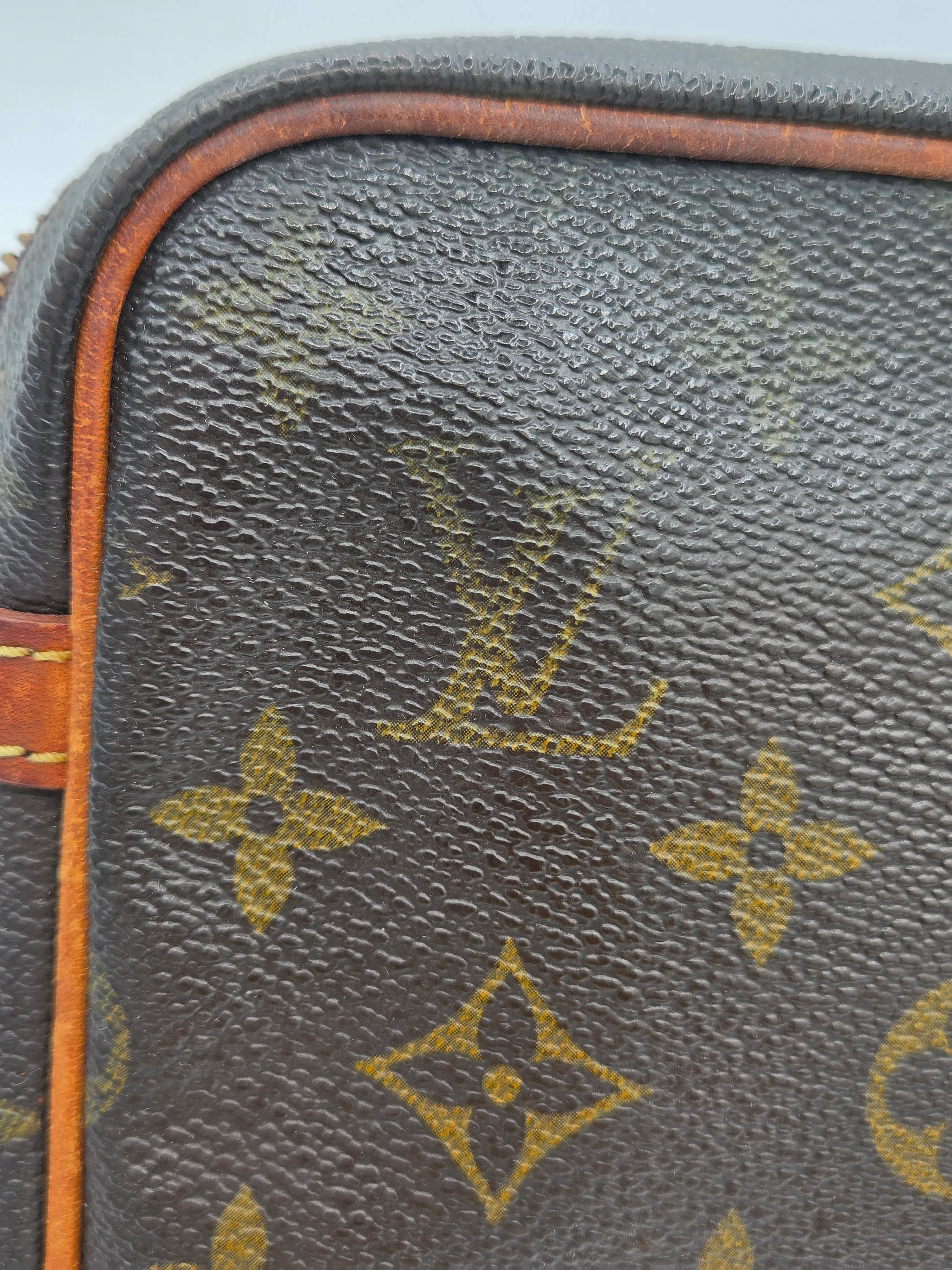 Close-up of a Louis Vuitton monogram canvas bag corner, showing the dark brown coated canvas with the classic tan/gold LV monogram pattern and a strip of orange-brown leather trim.