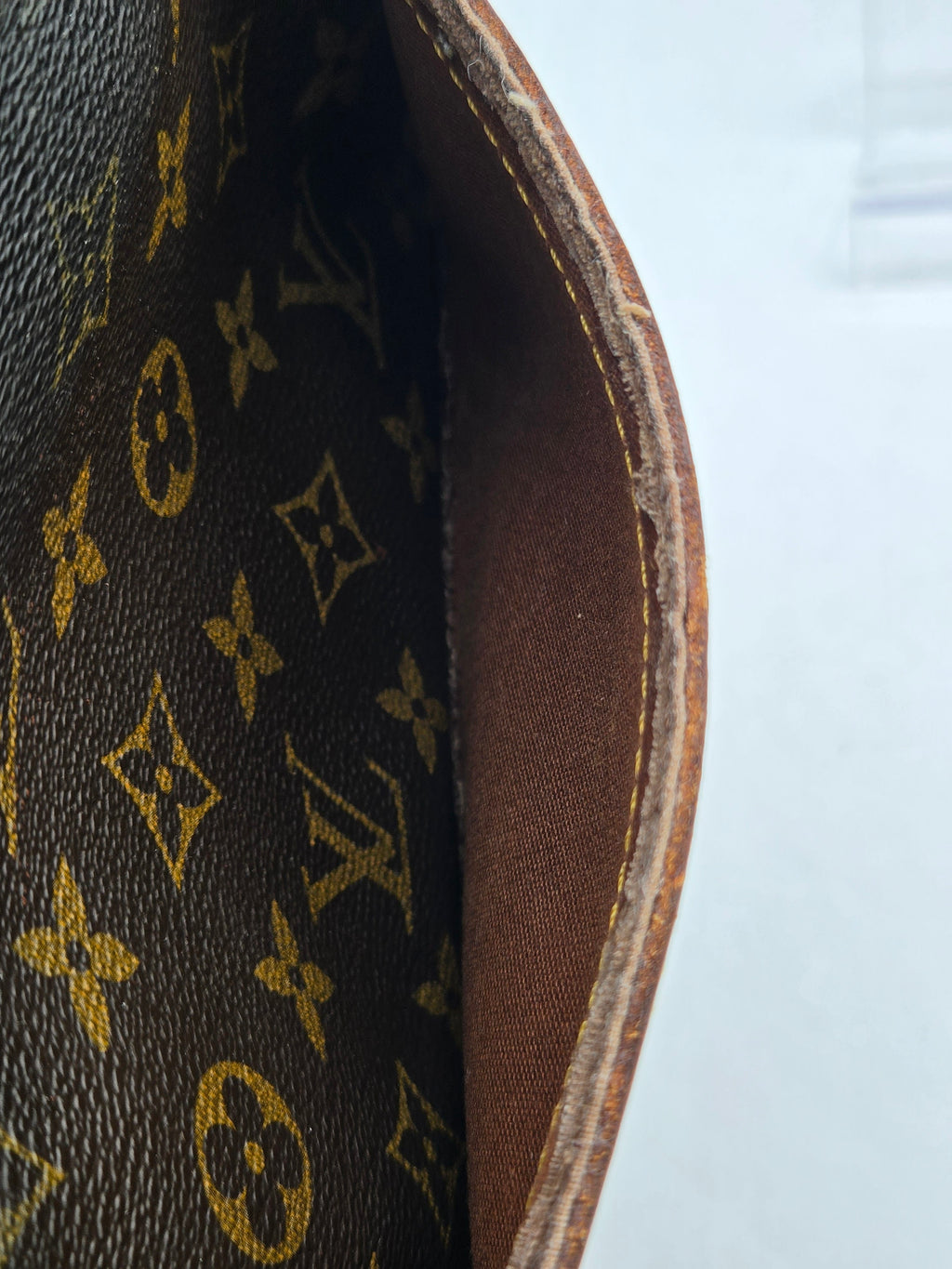 Close-up of a Louis Vuitton monogram canvas bag corner showing the dark brown coated canvas with gold LV monogram pattern and a brown leather edge with tan stitching.
