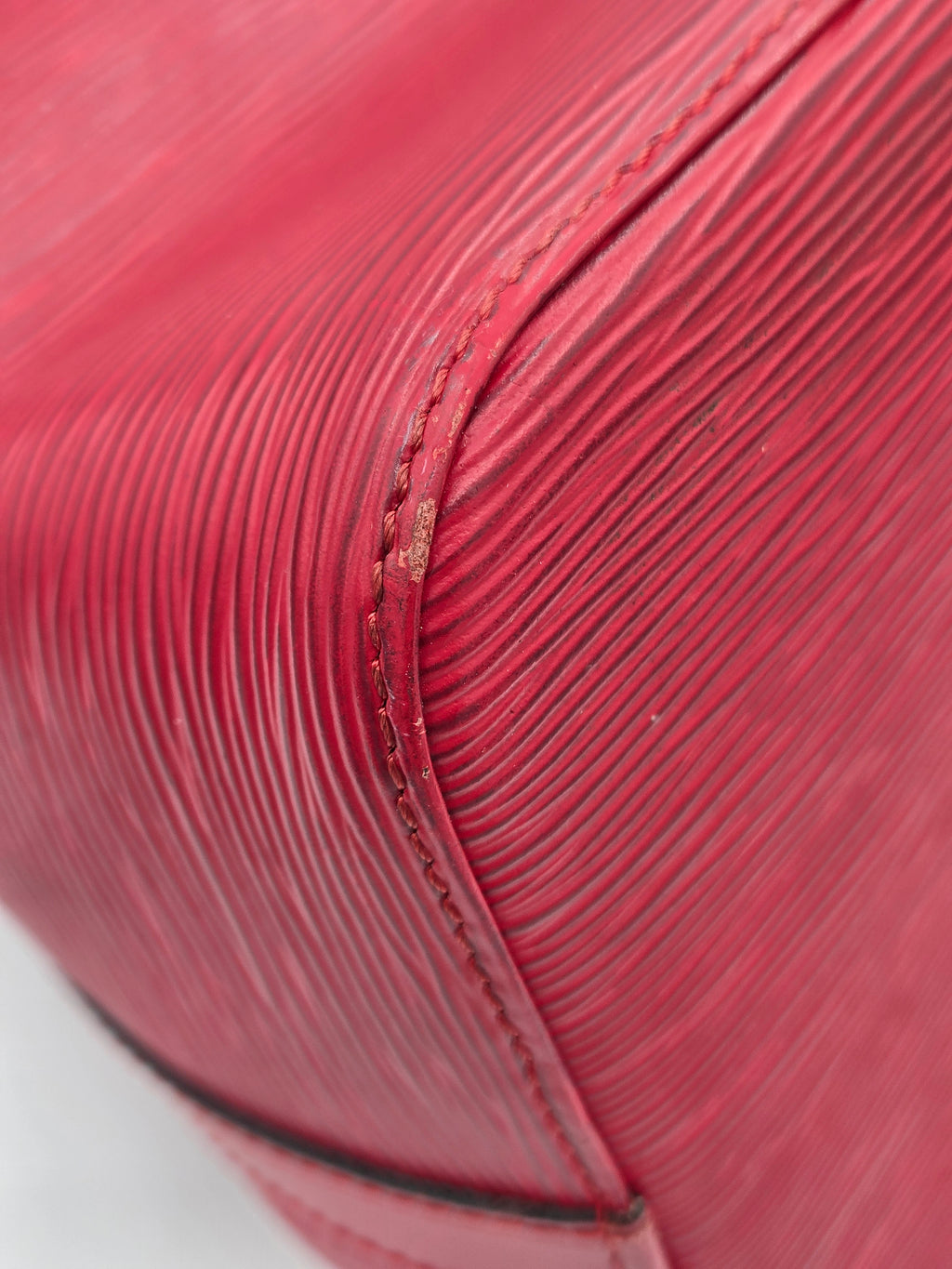 Close-up of a textured red leather handbag corner showing fine grain, stitching, and a dark edge trim against a white background.