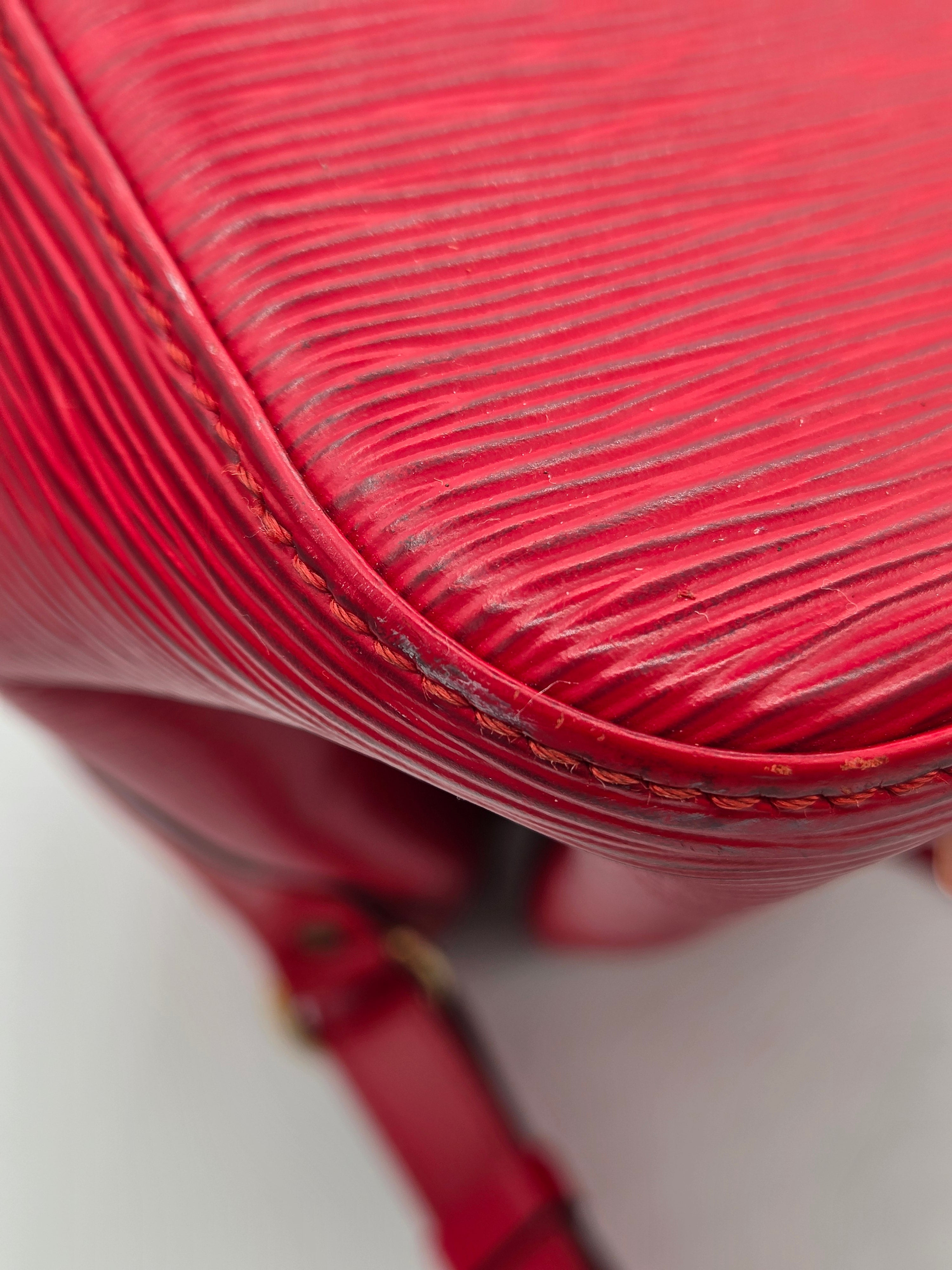 Close-up of a red textured leather handbag corner with visible stitching and a strap attachment, photographed against a light neutral background.