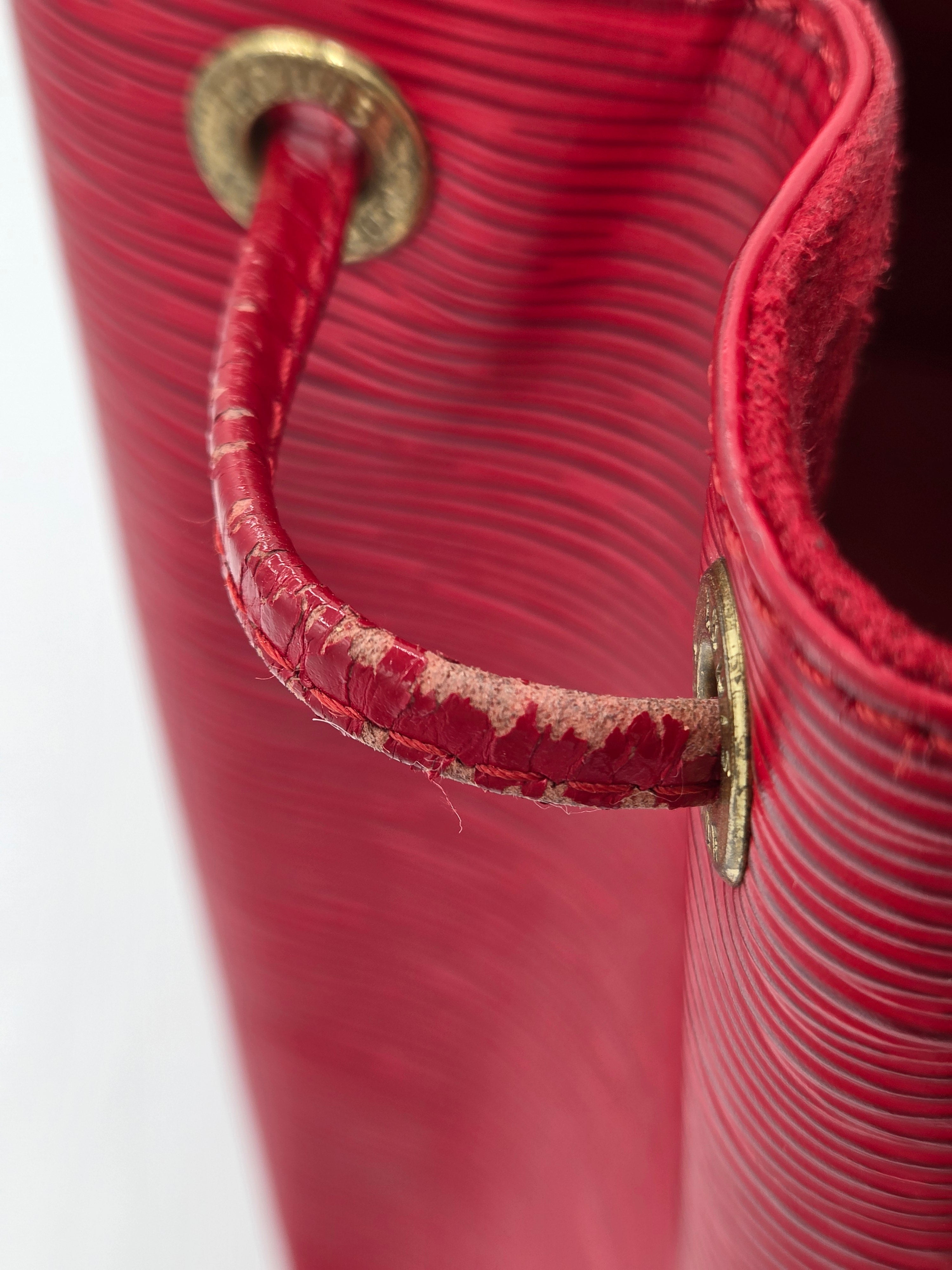 Close-up of a red textured leather handbag showing a curved leather strap with peeling distressed detailing and brass-tone metal grommets.