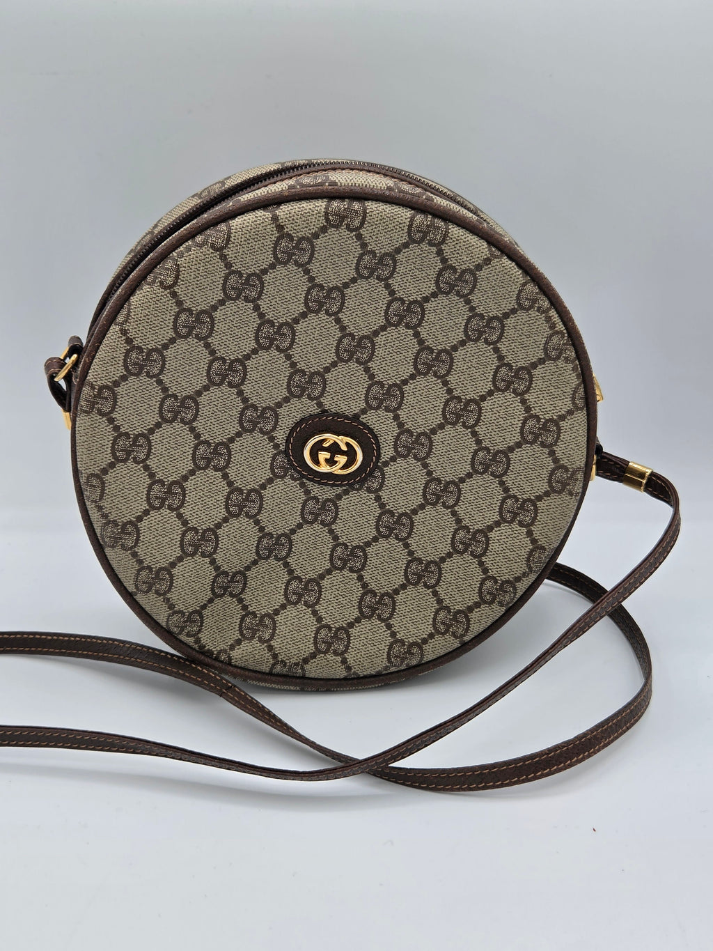 A round Gucci shoulder bag in the classic GG Supreme canvas, featuring a beige and brown monogram pattern, a central gold-tone GG logo clasp, and a matching brown leather strap.