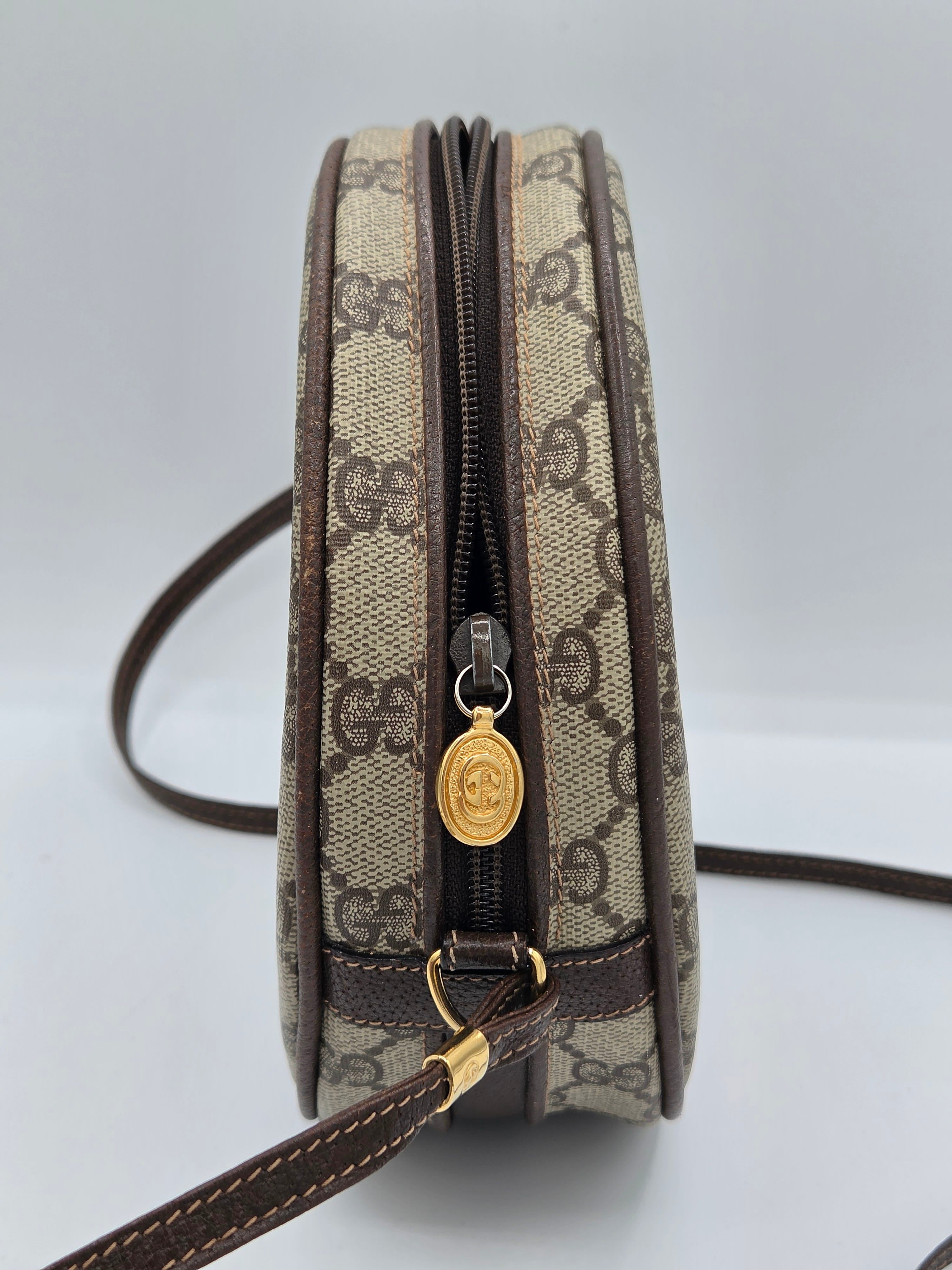 A Gucci monogram canvas crossbody bag shown from a side profile view, featuring the signature GG pattern in beige and brown with dark brown leather trim and a gold-tone interlocking CC charm on the zipper pull.