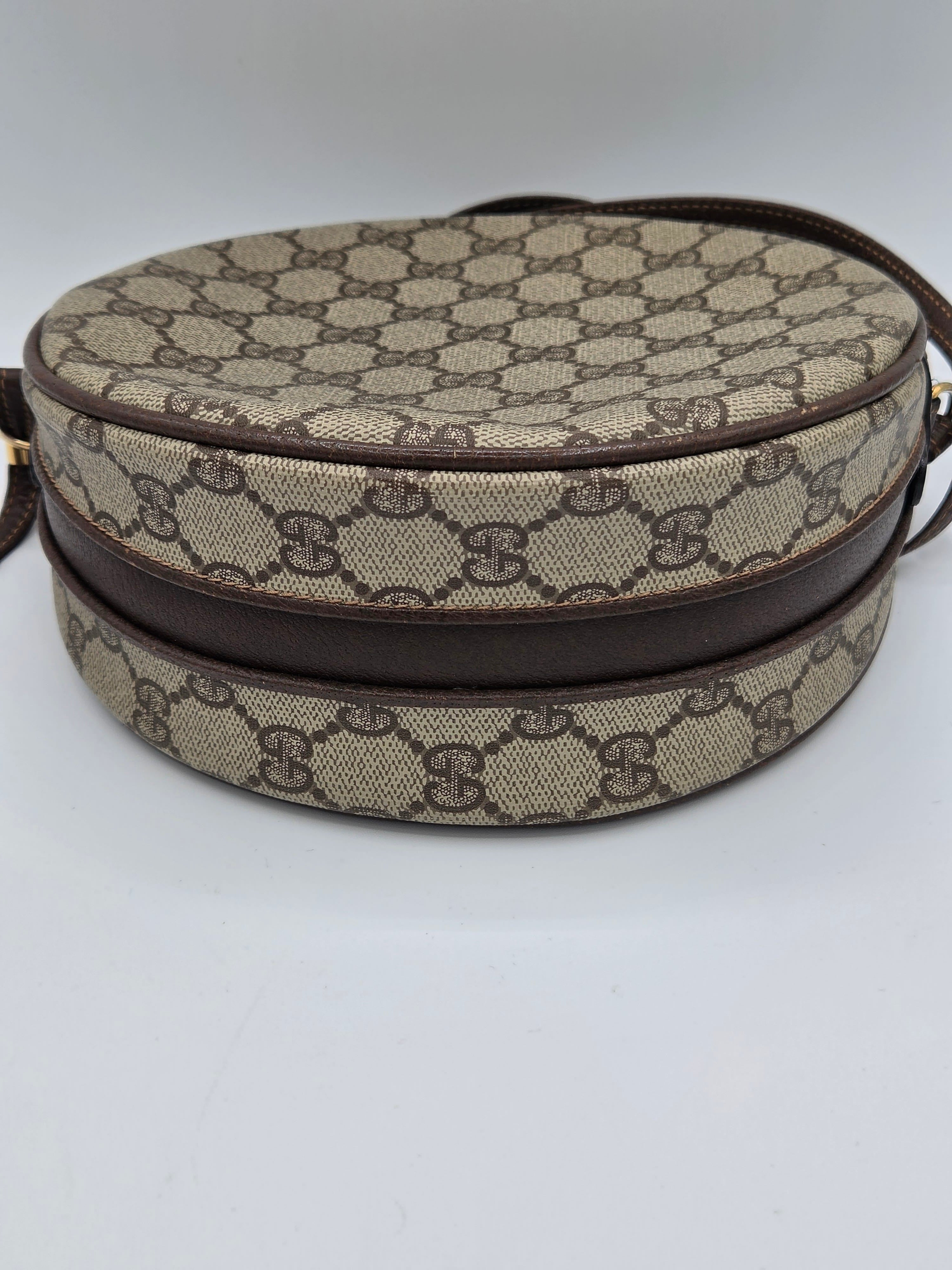 A round Gucci shoulder bag in beige and brown GG Supreme canvas with dark brown leather trim and a matching strap, shown on a light gray background.