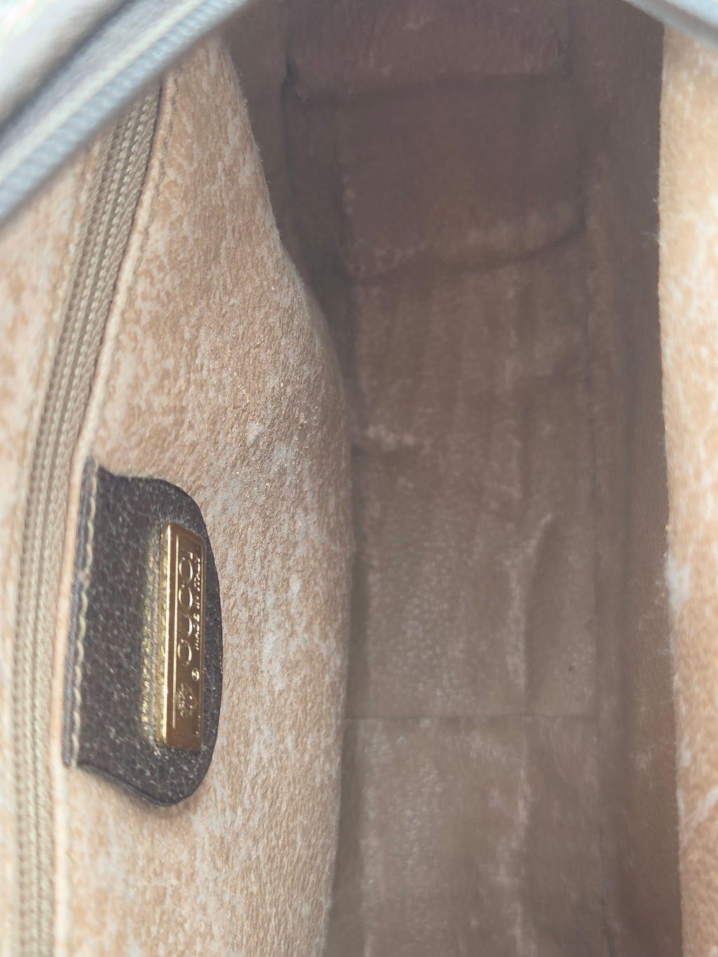 Close-up of the interior of a beige textured handbag, showing a zipper and a small gold-tone metal brand plaque on a dark tab.