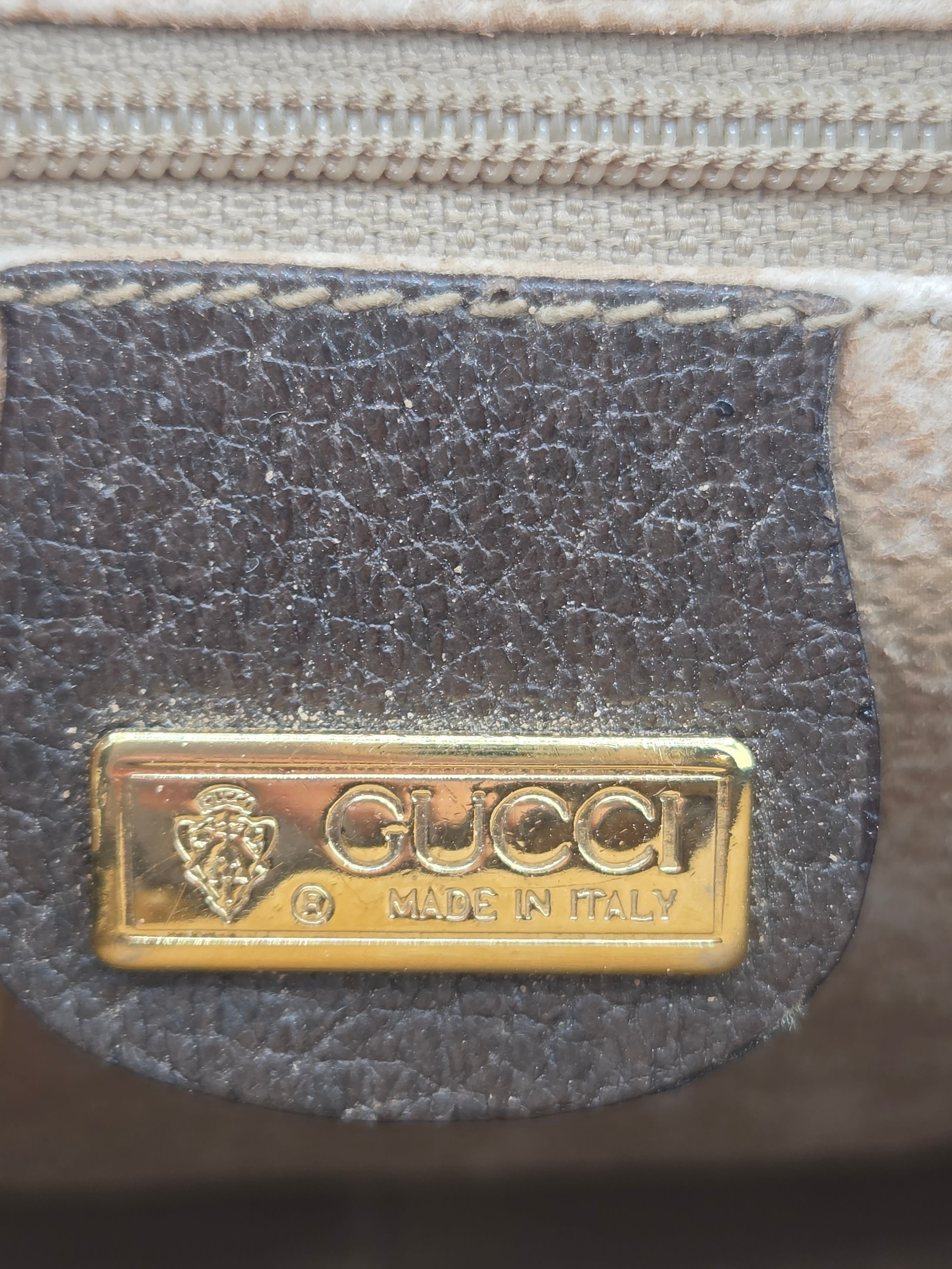 Close-up of a Gucci leather detail with a gold-tone metal plaque engraved with 'GUCCI MADE IN ITALY' and the brand's lion emblem, attached to a dark brown leather tab on a beige woven textile.