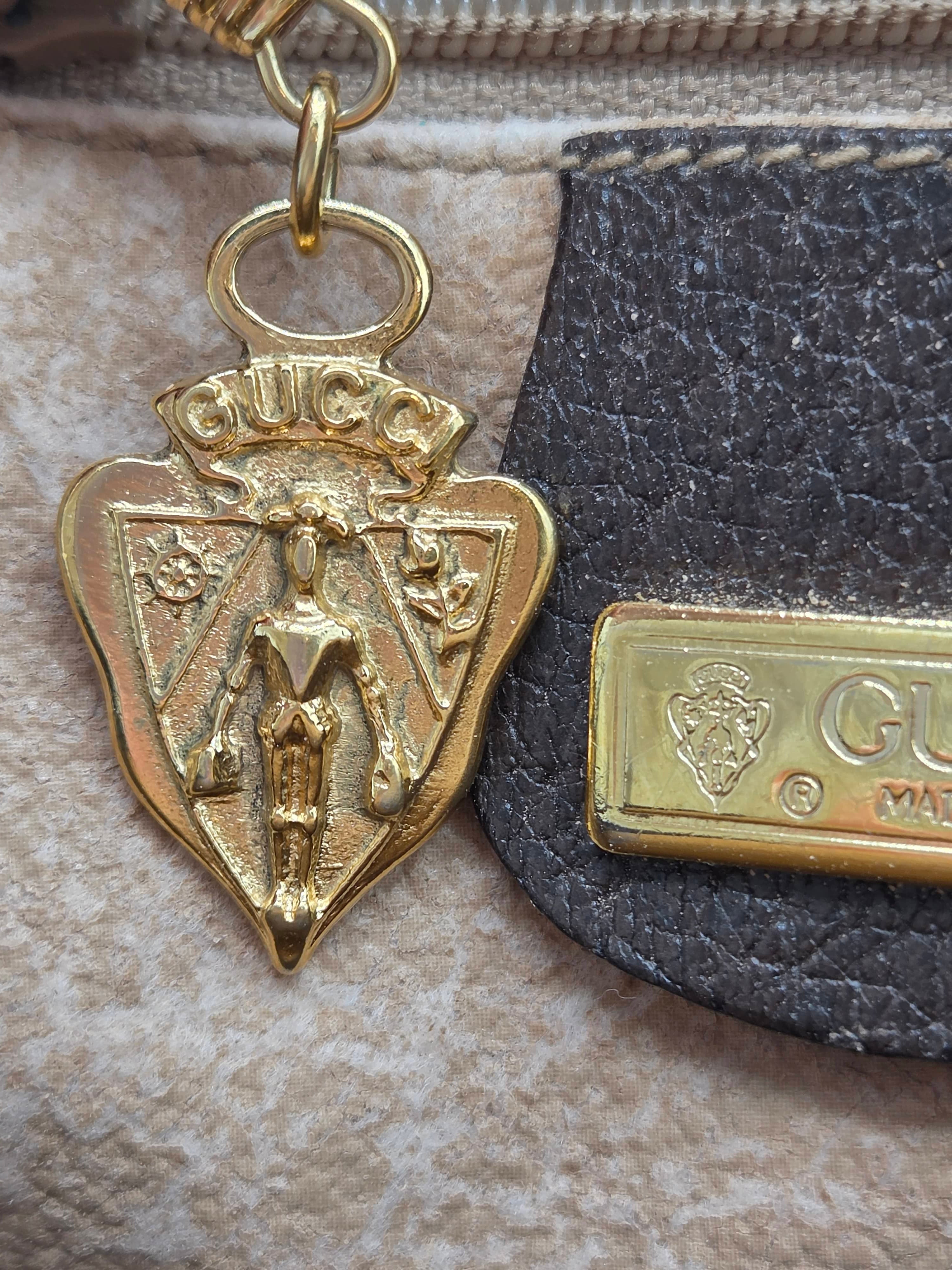 Close-up of a Gucci accessory featuring a gold-tone shield-shaped emblem with a crowned lion motif, attached to a dark textured strap next to a gold-tone Gucci bar, on a beige textured fabric background.