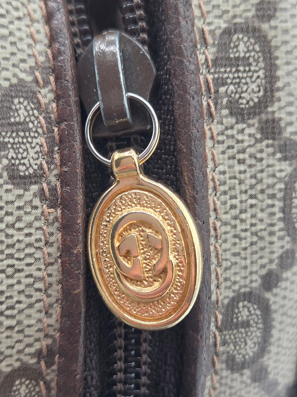 Close-up of a Gucci zippered pouch featuring the brand's signature GG monogram canvas, dark brown leather trim, and a gold-tone oval interlocking G logo zipper pull.