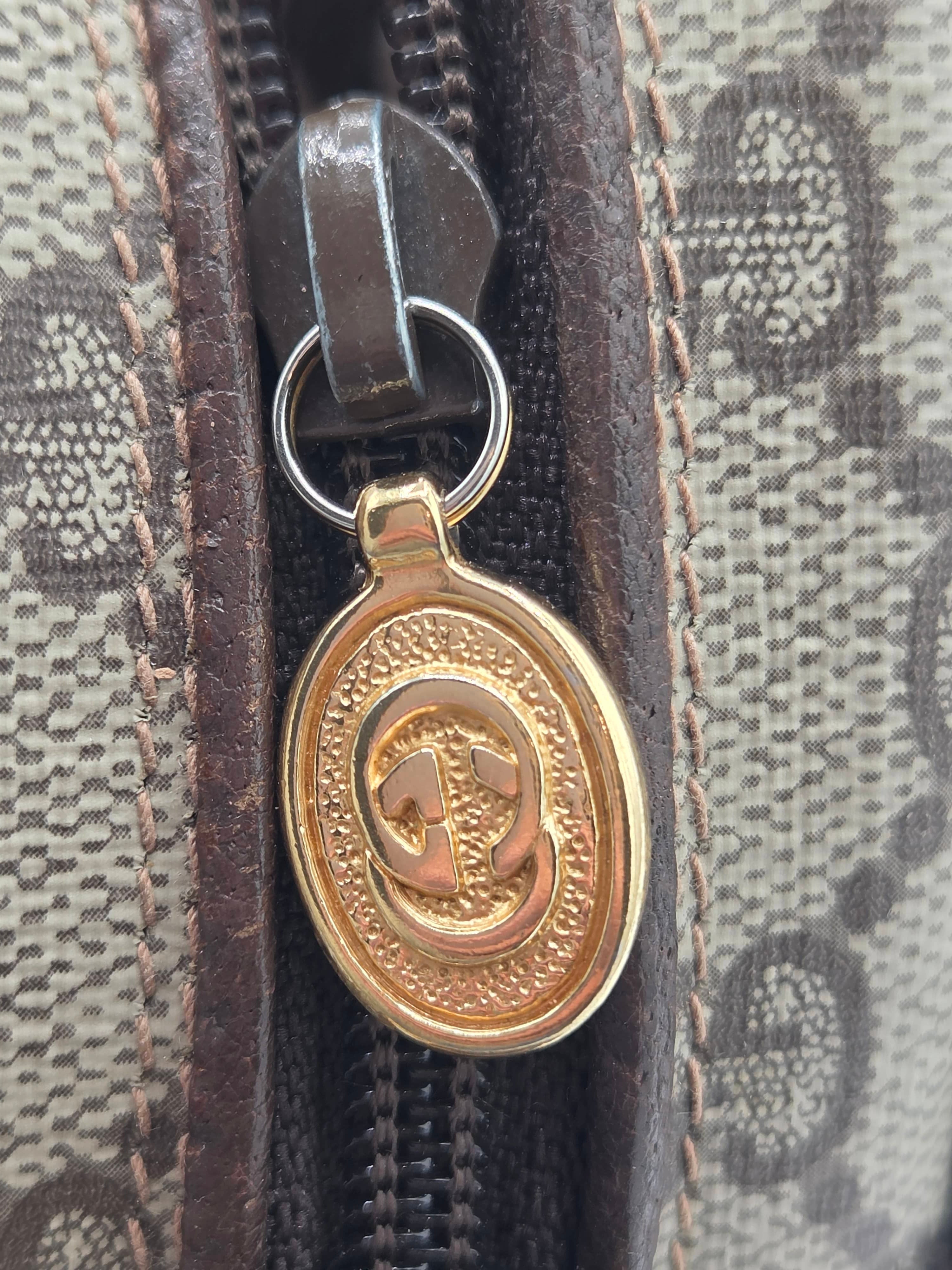 Close-up of a Gucci zippered pouch featuring the brand's signature GG monogram canvas, dark brown leather trim, and a gold-tone oval interlocking G logo zipper pull.
