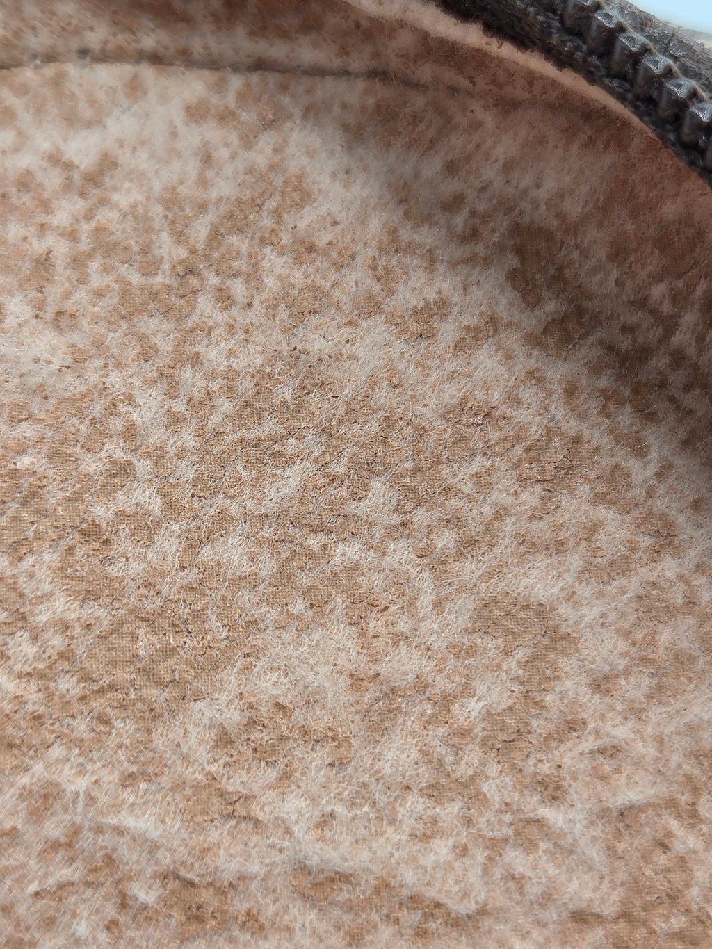 Close-up of a textured, looped fabric or felt material in a warm tan-brown color, showing the dense fibrous surface and a dark zipper edge at the top right.
