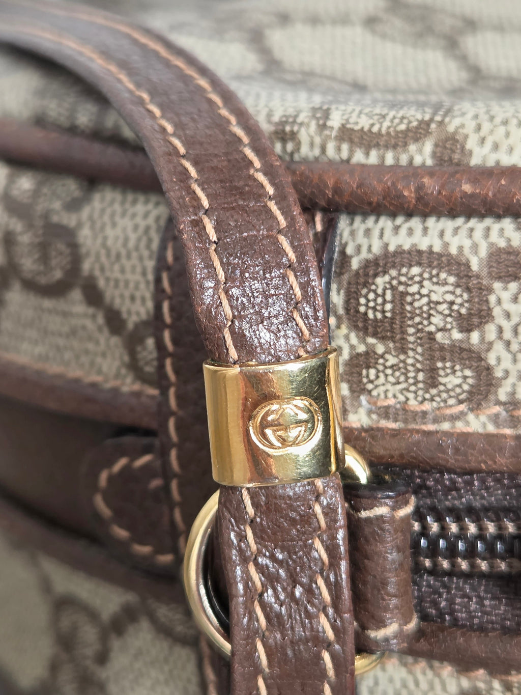 Close-up of a Gucci handbag featuring the brand's classic GG monogram canvas in beige and brown, a brown leather strap with beige contrast stitching, and a gold-tone metal clasp with the interlocking G logo.