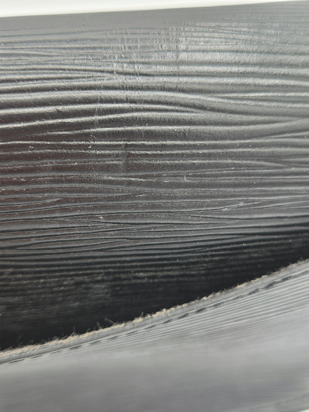Close-up of a dark gray or black textured material with horizontal ridges and a stitched edge visible in the lower foreground.