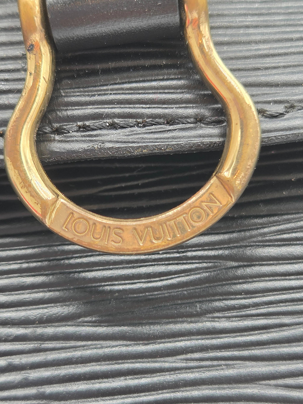 Close-up of a Louis Vuitton handbag featuring a textured black leather strap and a gold-tone metal D-ring with the brand name engraved.