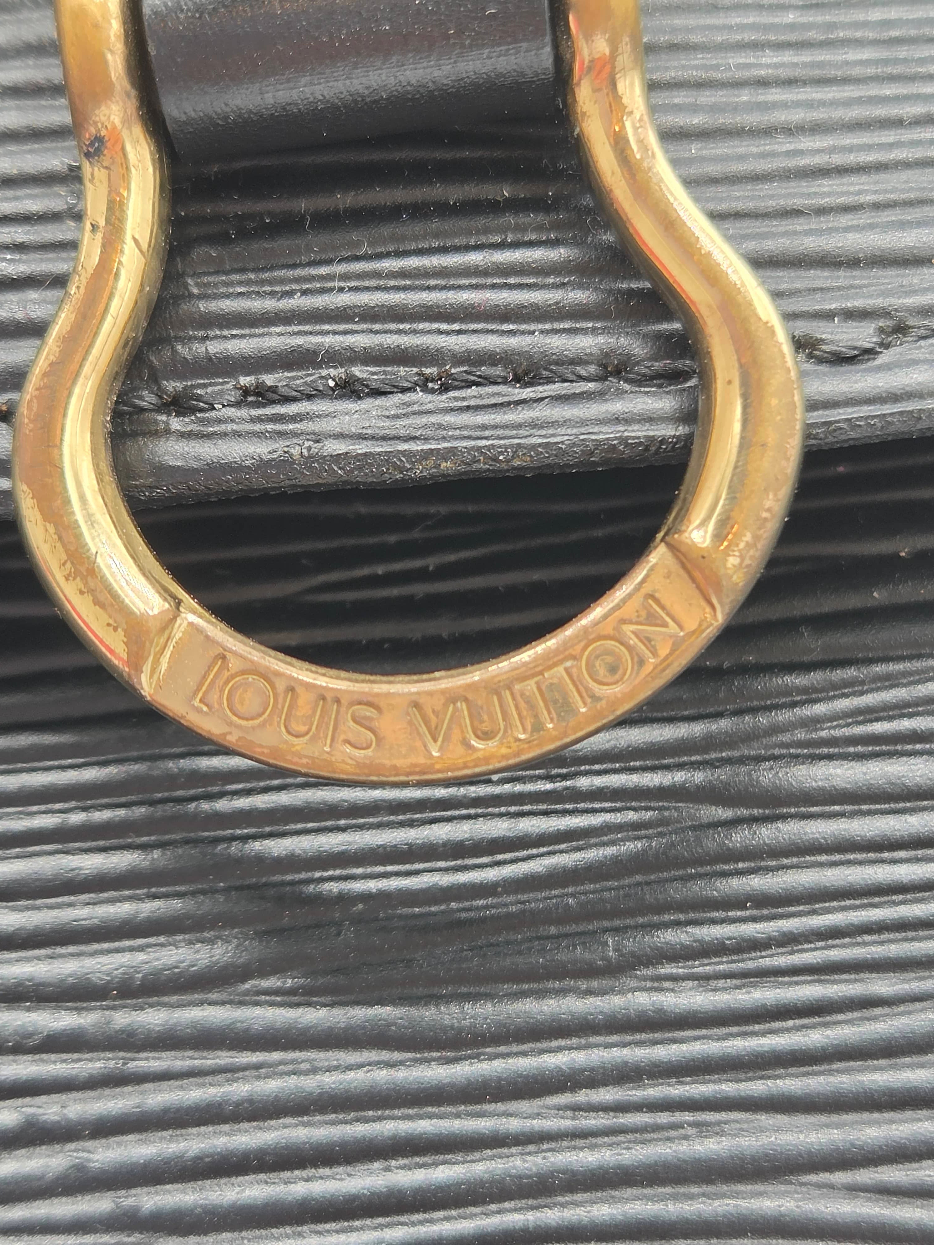 Close-up of a Louis Vuitton handbag featuring a textured black leather strap and a gold-tone metal D-ring with the brand name engraved.