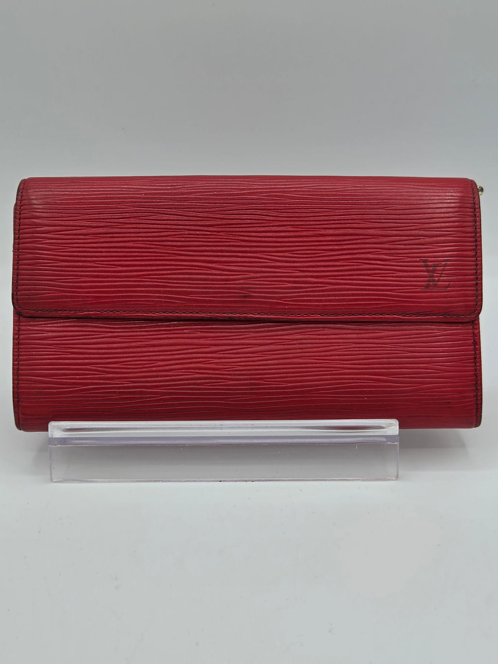 A red textured leather wallet or clutch with a vertical grain pattern, shown closed with a clear plastic sleeve partially inserted on the right side, set against a plain light gray background.