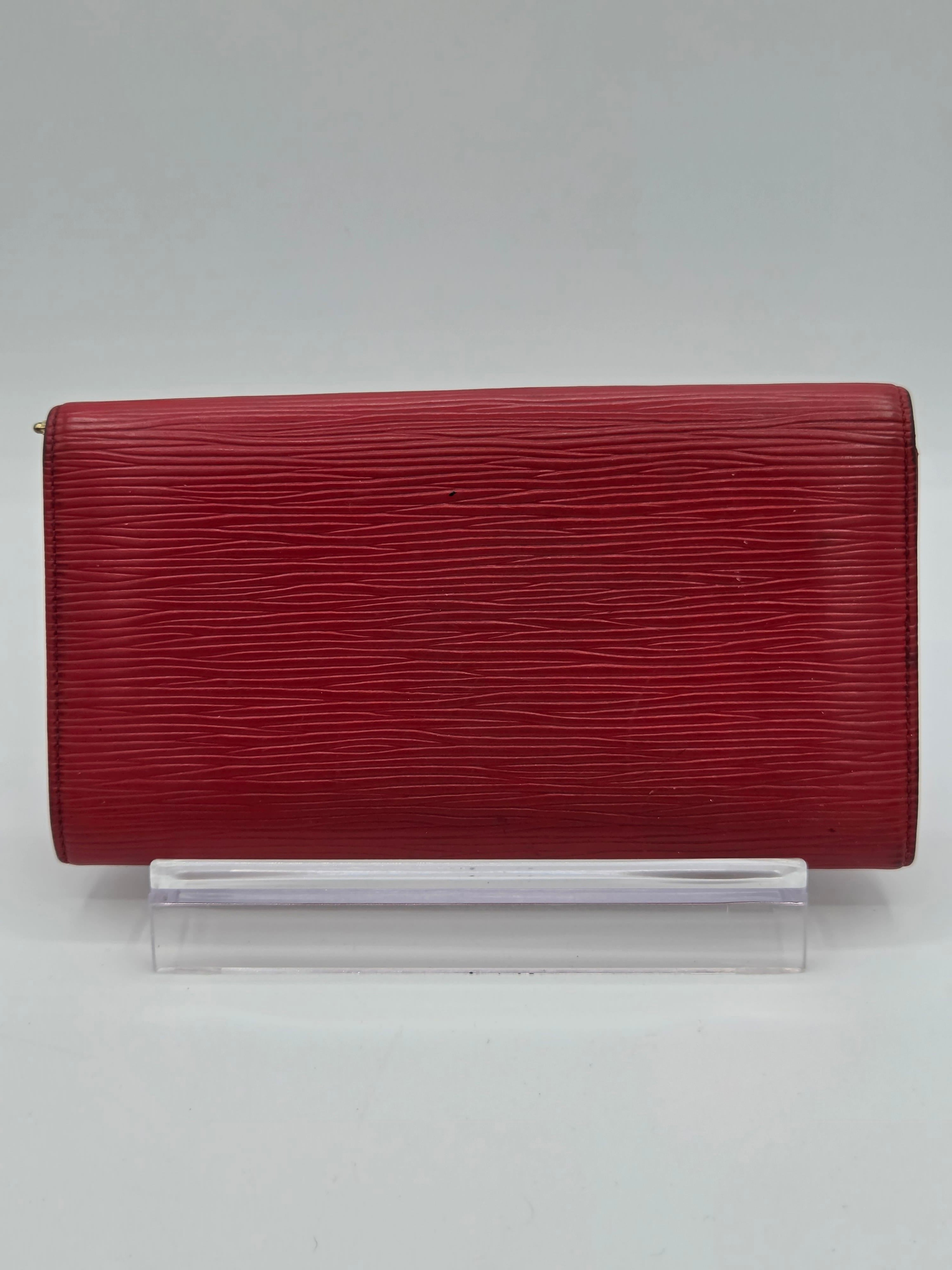 A red textured leather clutch or wallet with a vertical grain pattern, shown closed next to a clear plastic sleeve on a light gray background.