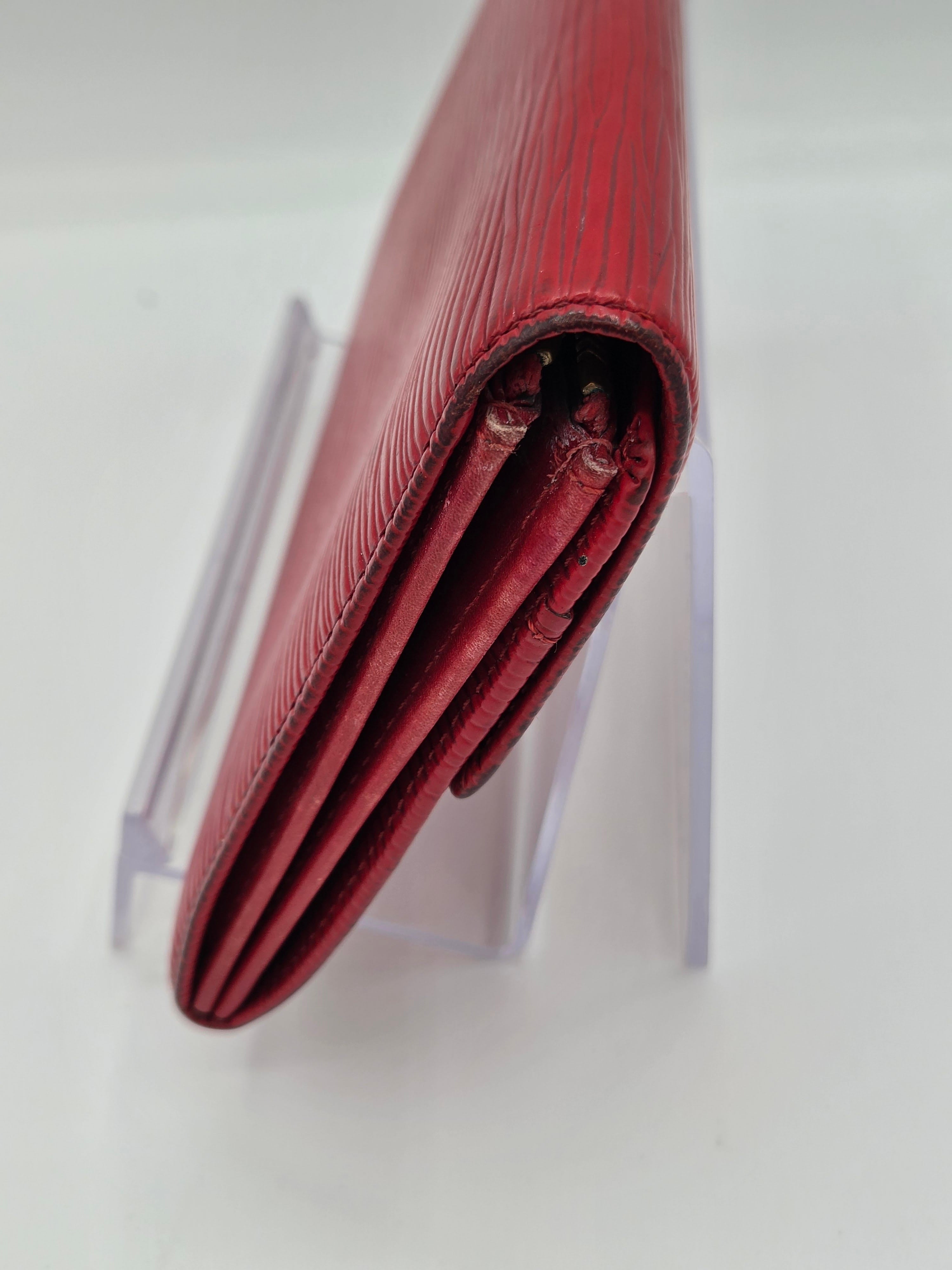 A close-up of a folded red leather wallet or clutch with a textured pattern, resting on a clear acrylic display stand against a white background.