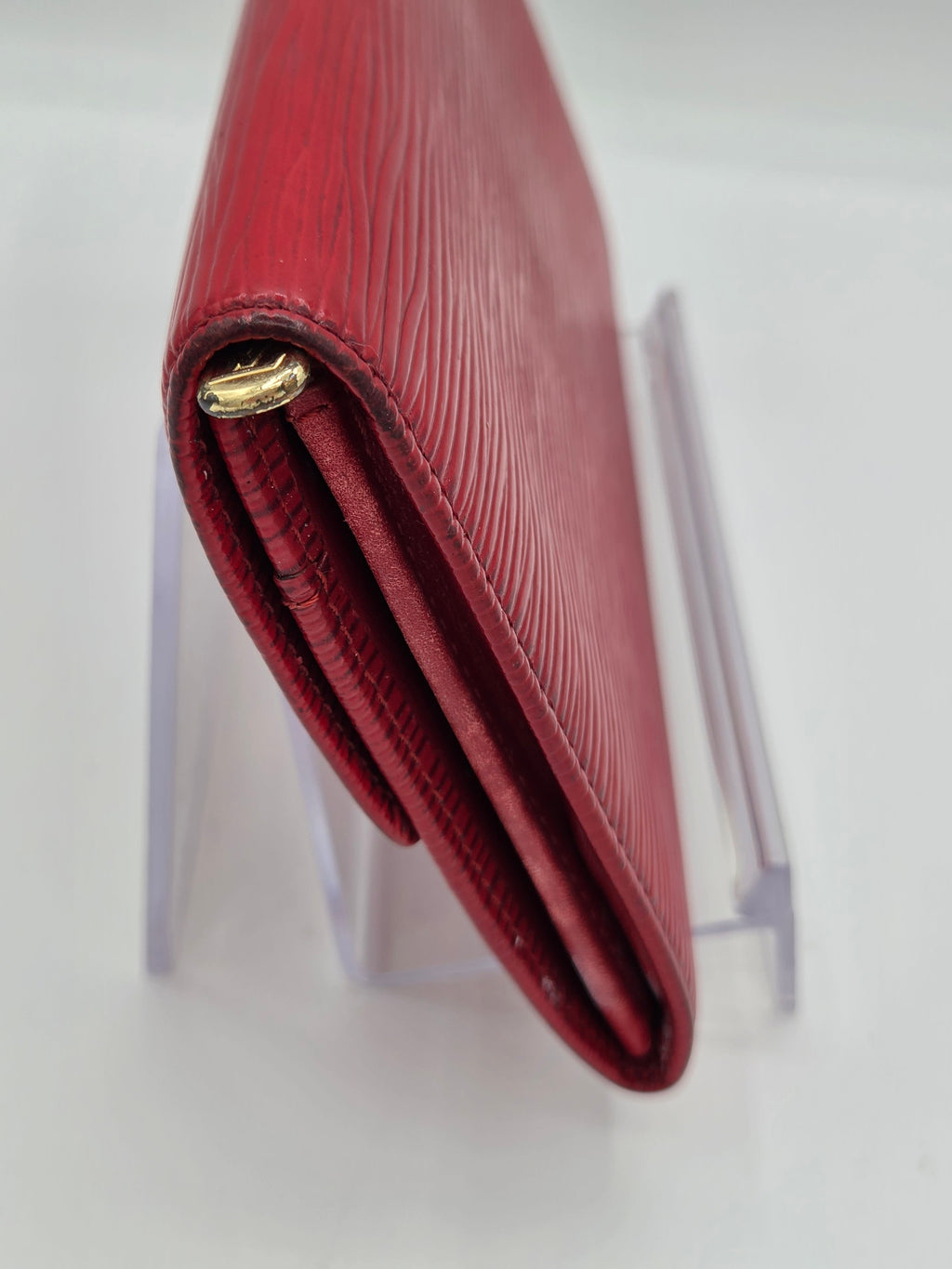 A close-up of a red textured leather wallet with a gold-tone metal clasp, resting on a clear acrylic display stand against a white background.