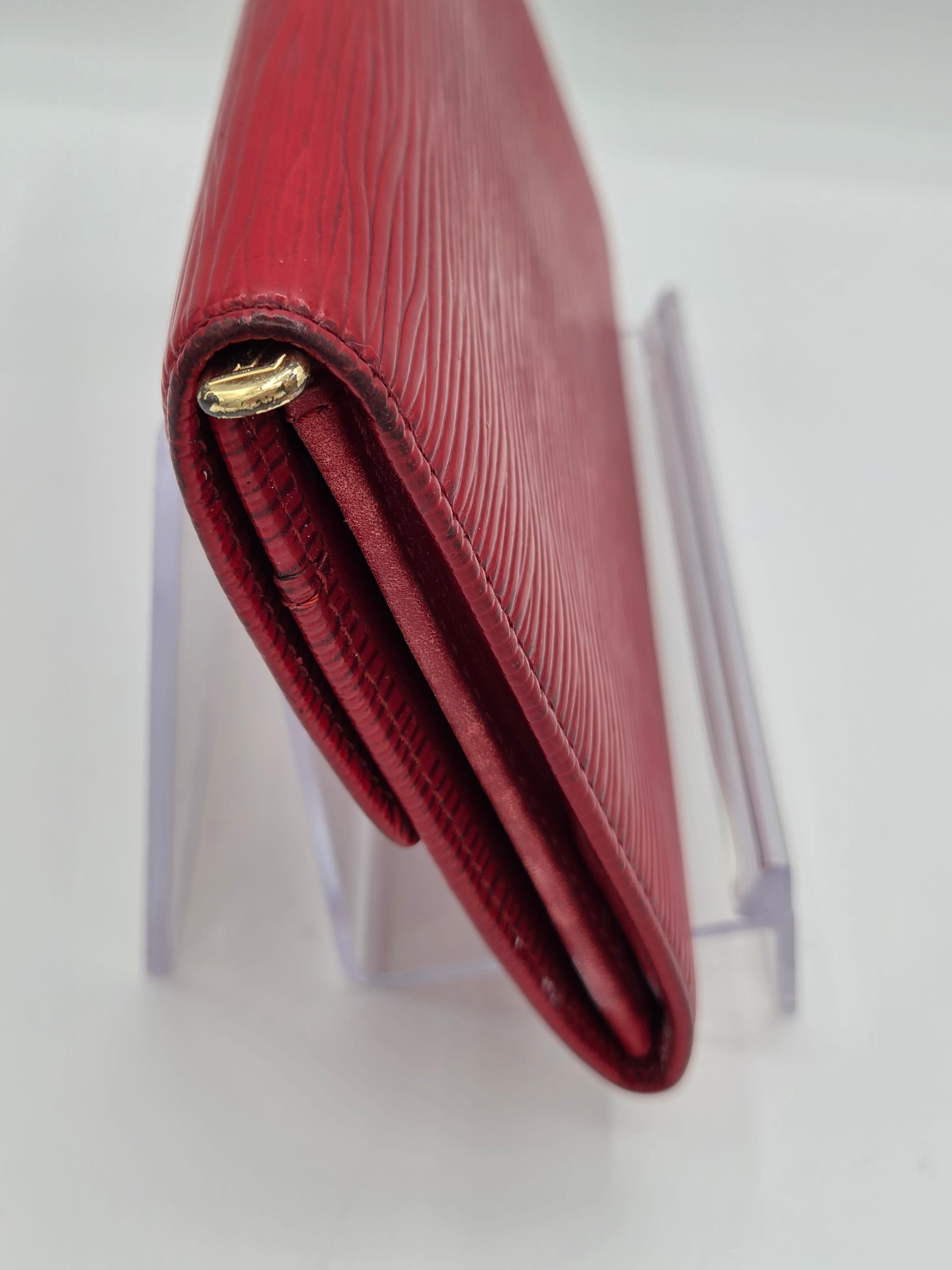 A close-up of a red textured leather wallet with a gold-tone metal clasp, resting on a clear acrylic display stand against a white background.