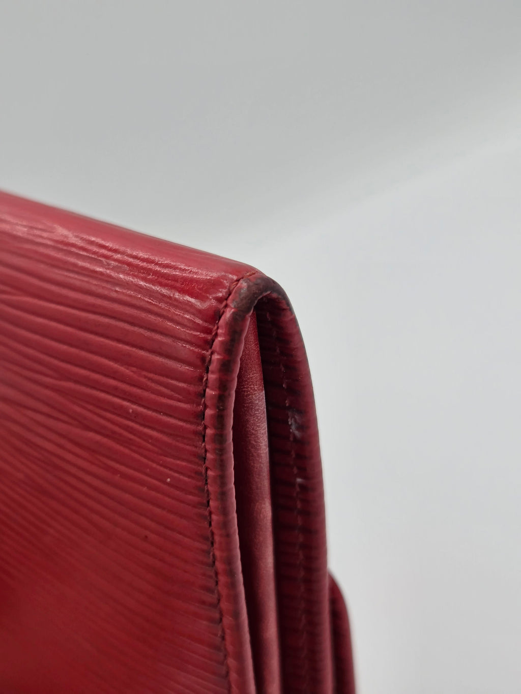 Close-up of a red textured leather wallet corner showing the fine grain and stitching against a light gray background.