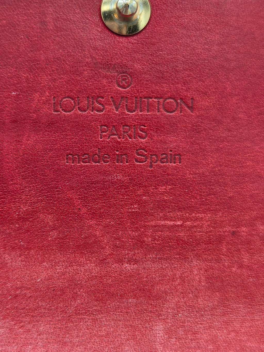 Close-up of a red textured leather surface with embossed Louis Vuitton Paris made in Spain text and a small metallic snap on the left edge.