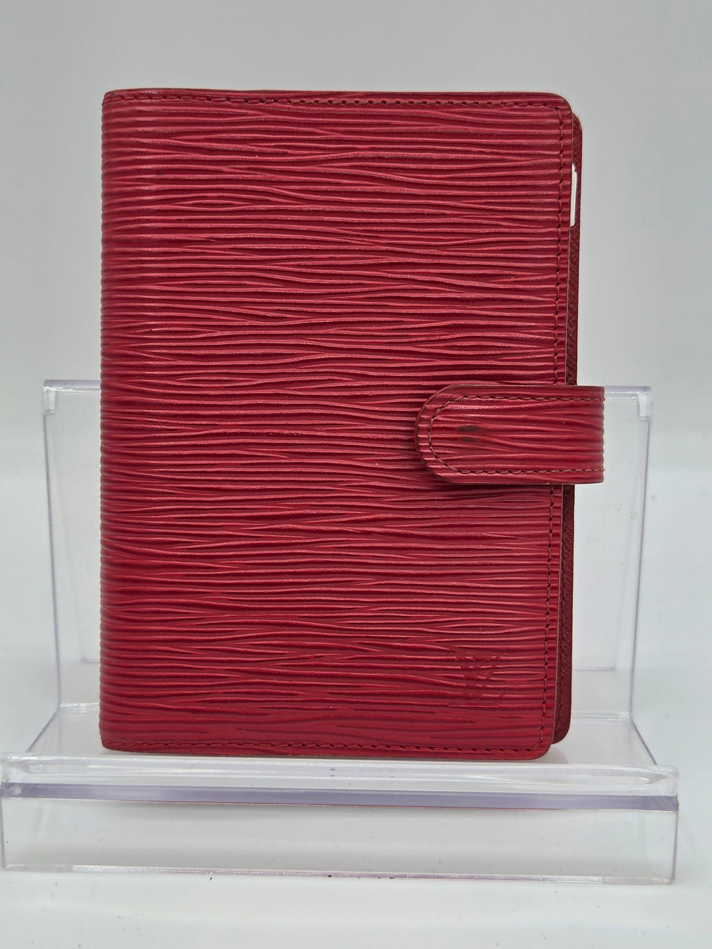 A red textured leather wallet or clutch with a vertical grain pattern and a top strap closure, displayed on a clear acrylic stand against a light gray background.