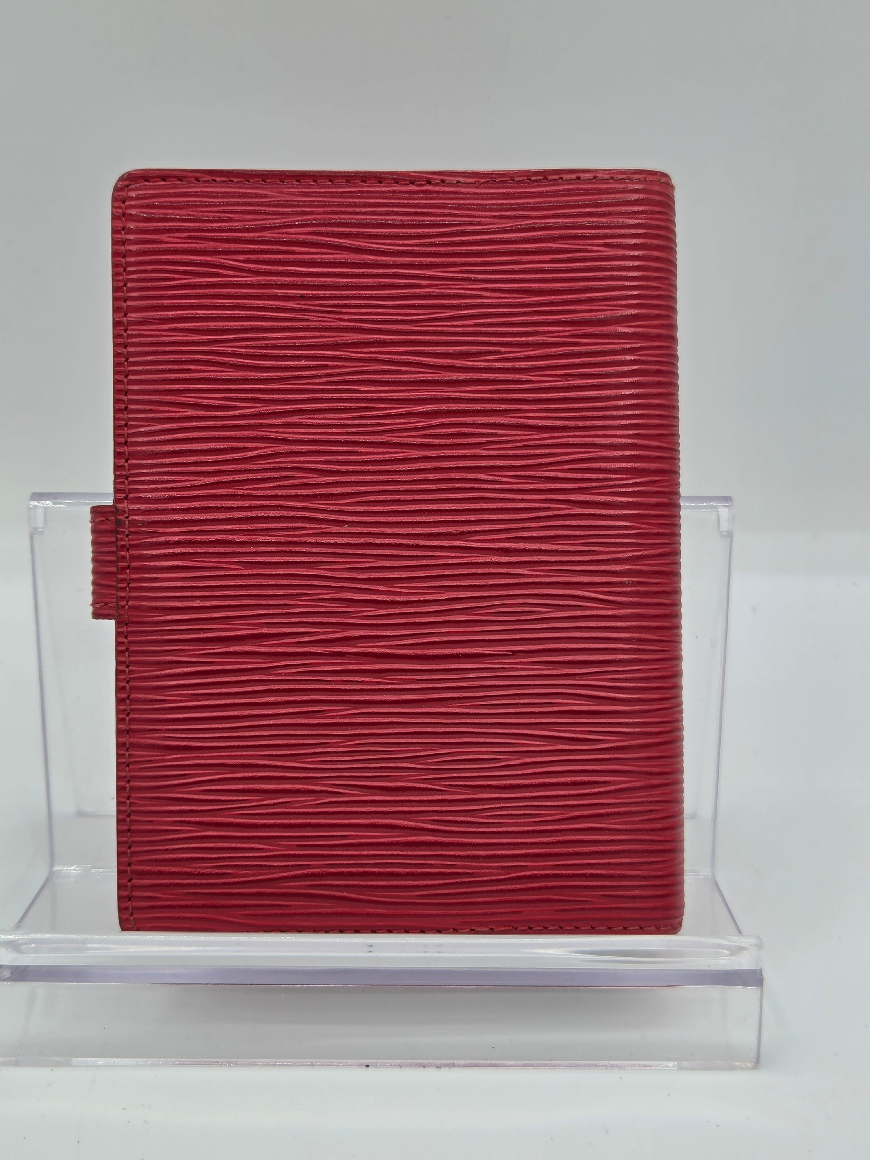 A red textured leather clutch or wallet with a vertical grain pattern, displayed on a clear acrylic stand against a plain white background.