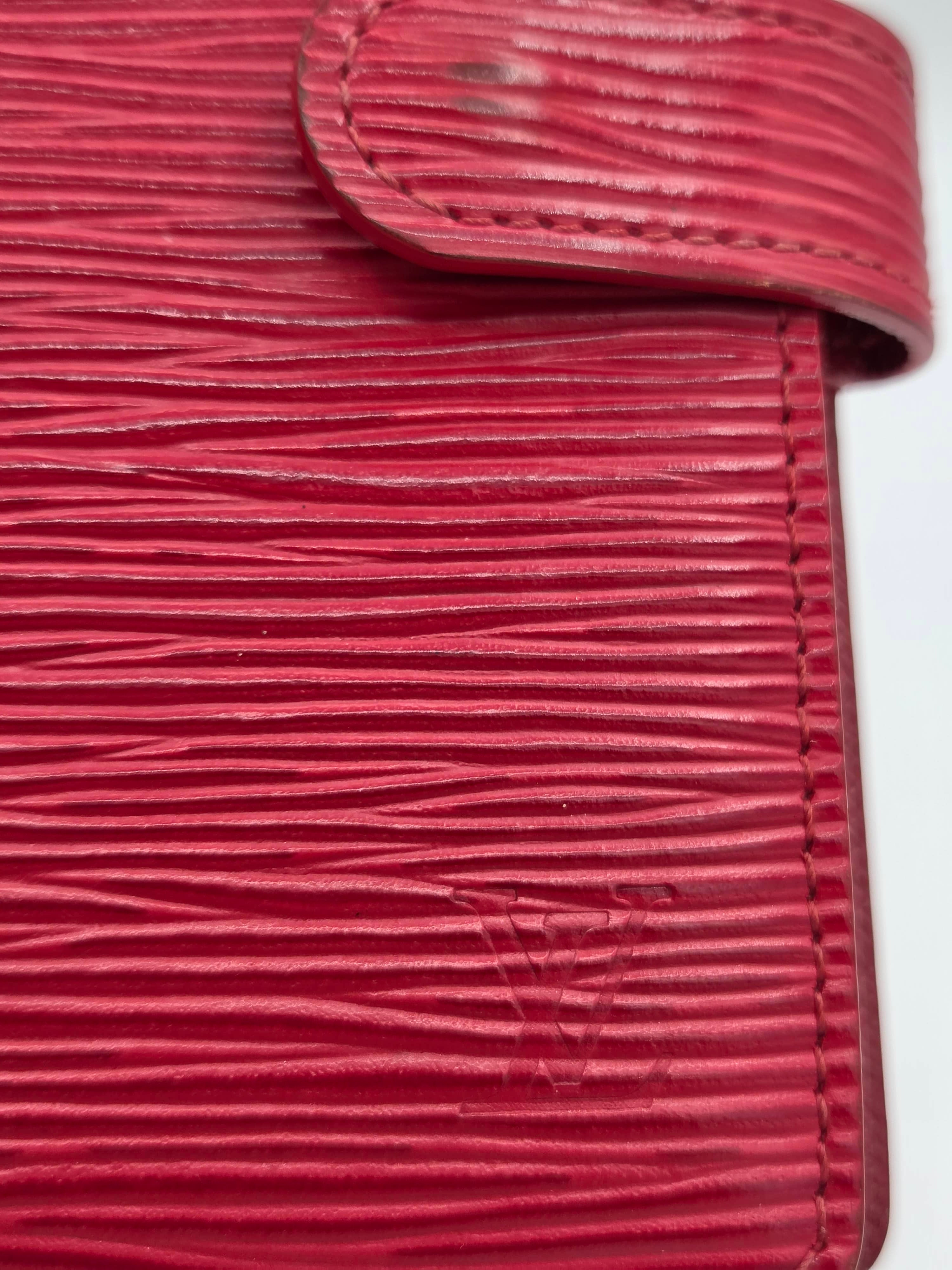 A close-up of a red textured leather wallet or card holder with a snap tab closure and subtle embossed Louis Vuitton monogram.