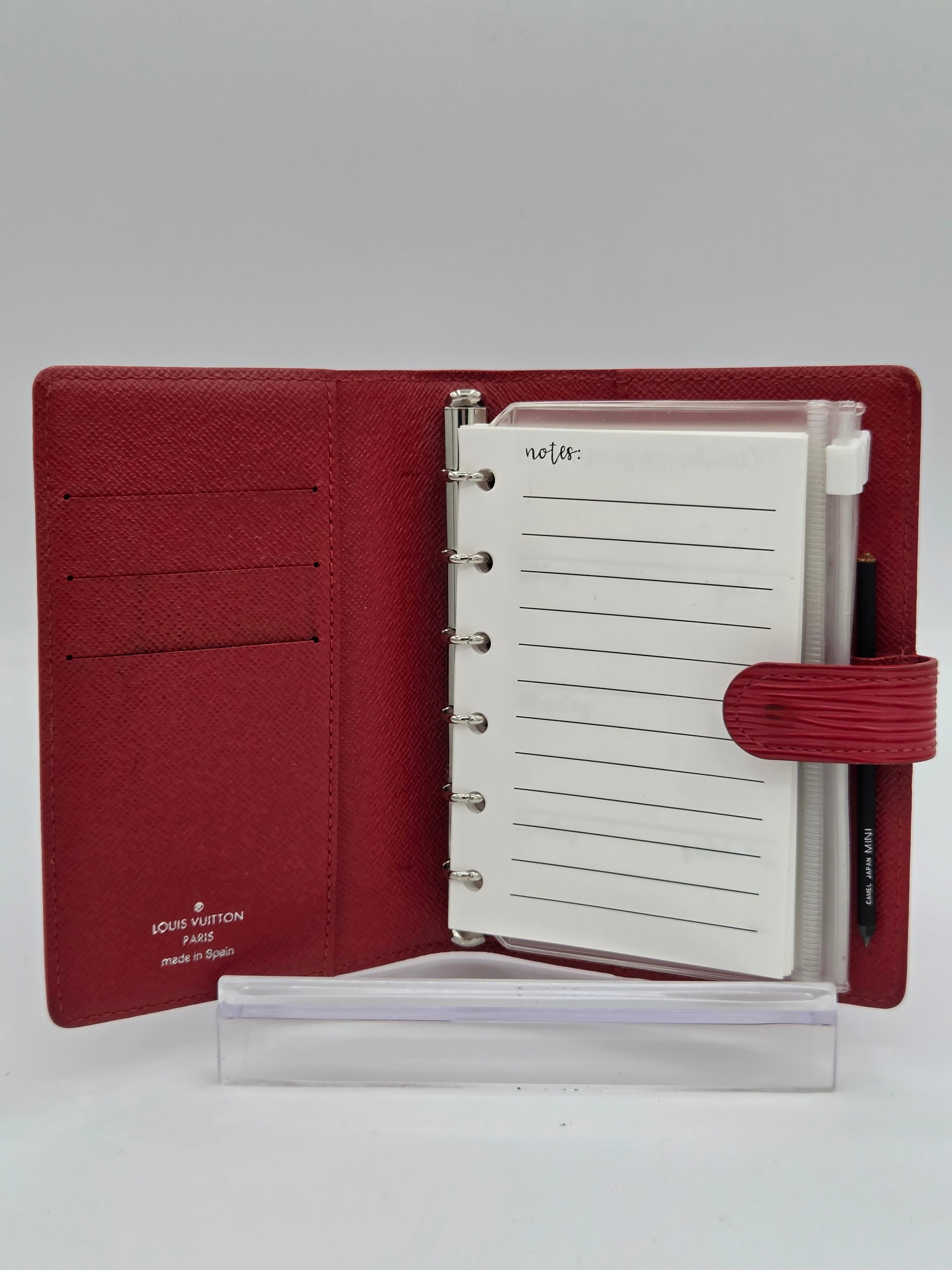 A Louis Vuitton red leather planner shown open, featuring card slots on the left, lined notepad pages in the center, and a pen loop with a pen on the right, displayed on a clear acrylic stand against a neutral gray background.