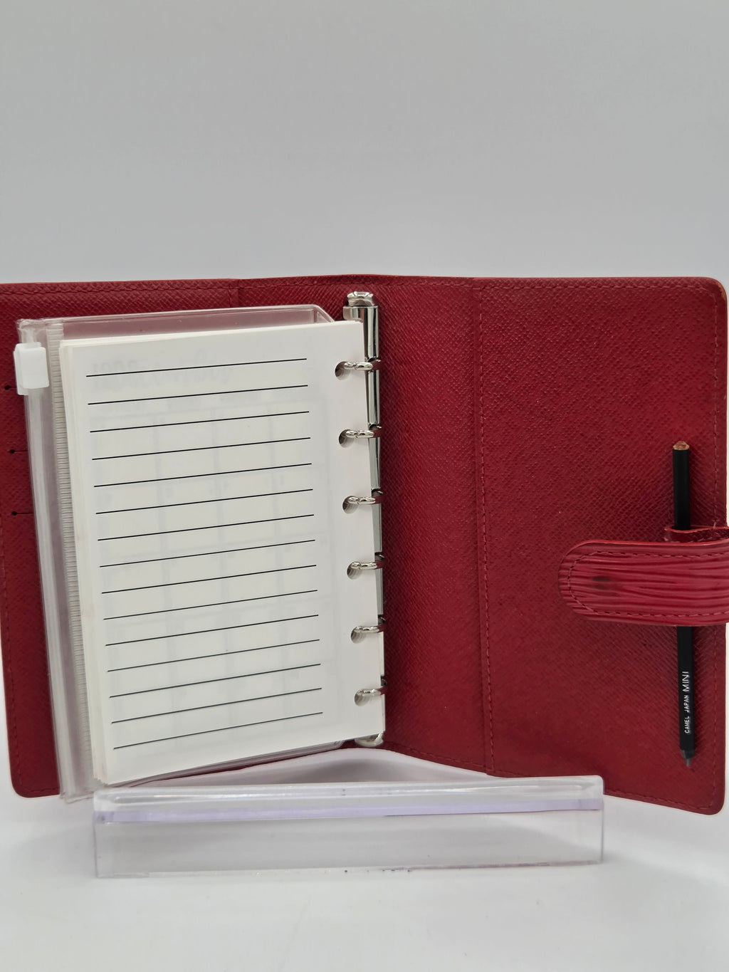 A red textured ring-bound planner or notebook cover shown open, with a white lined page inserted, a black pen in the top loop, and a clear plastic sleeve standing beside it on a light gray background.