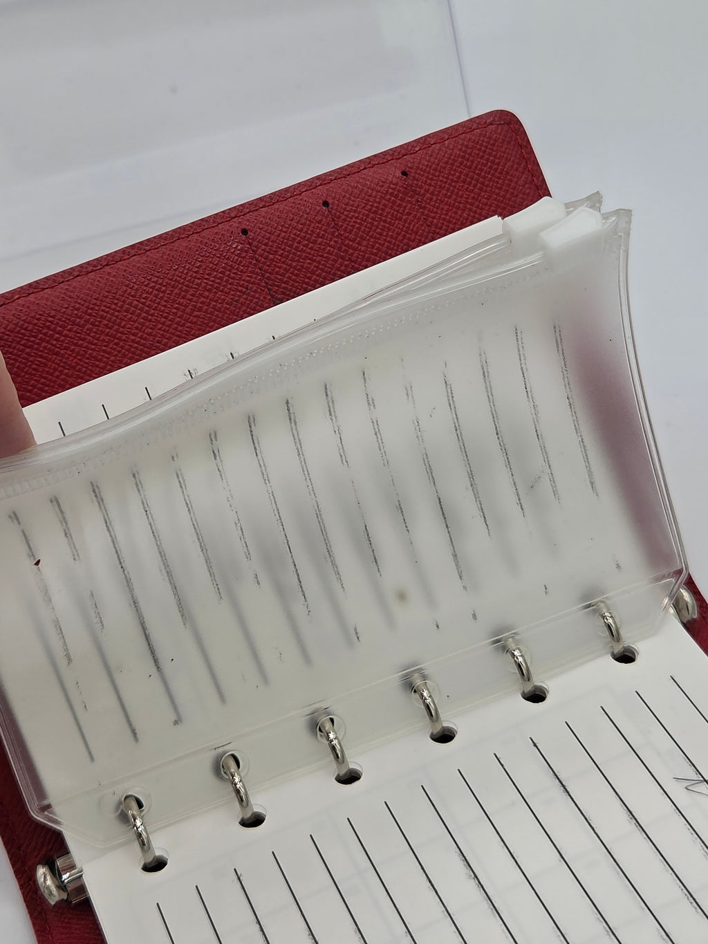 A close-up of a red textured ring binder notebook with clear plastic pockets holding white lined paper inserts, shown against a light background.