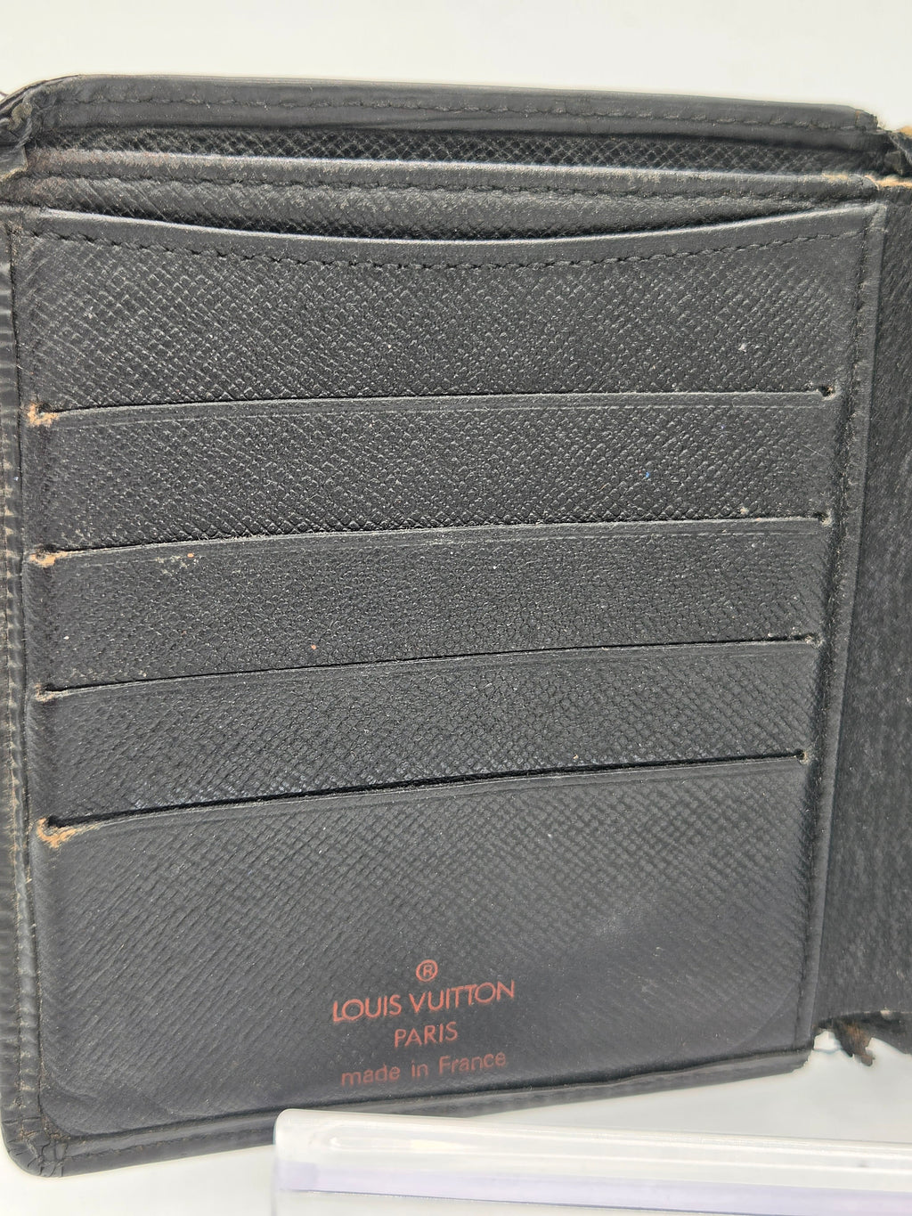 A close-up of a black Louis Vuitton bi-fold wallet made of textured Saffiano leather, showing the interior card slots and the brand's logo printed in red on the bottom left.