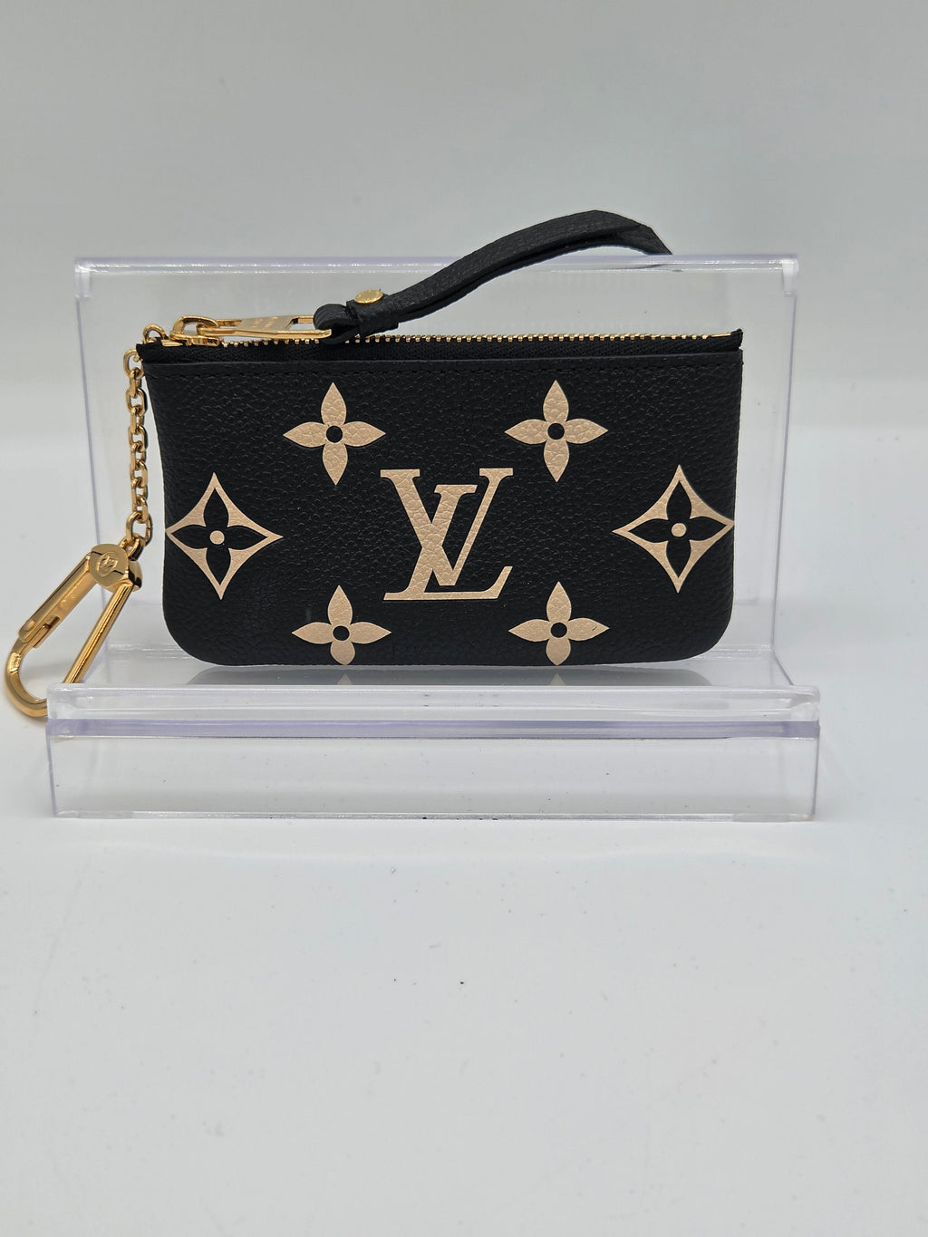 A Louis Vuitton black monogram wristlet pouch with gold-tone hardware, displayed inside a clear acrylic stand on a light gray background.